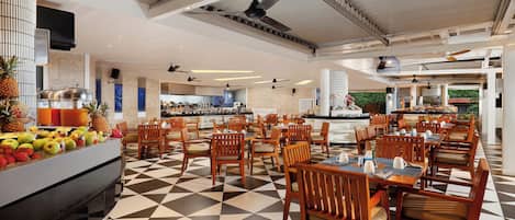 Een restaurantgedeelte van een hotel met een geruit vloertapijt, houten stoelen en een buffet met fruit.