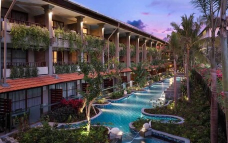 A hotel with multiple balconies, a central swimming pool, and lush landscaping.