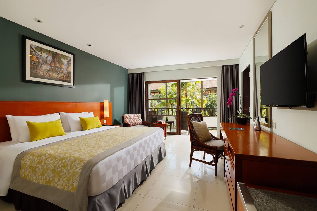 Een hotelkamer met een groot bed, een bureau, een televisie en een balkon met uitzicht op groen.