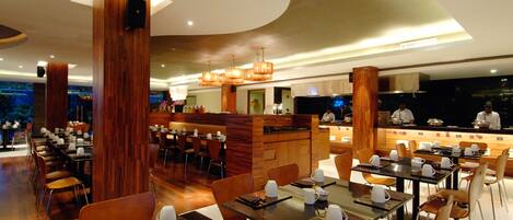 A restaurant with multiple dining tables and chairs, wooden pillars, and a kitchen area in the background.