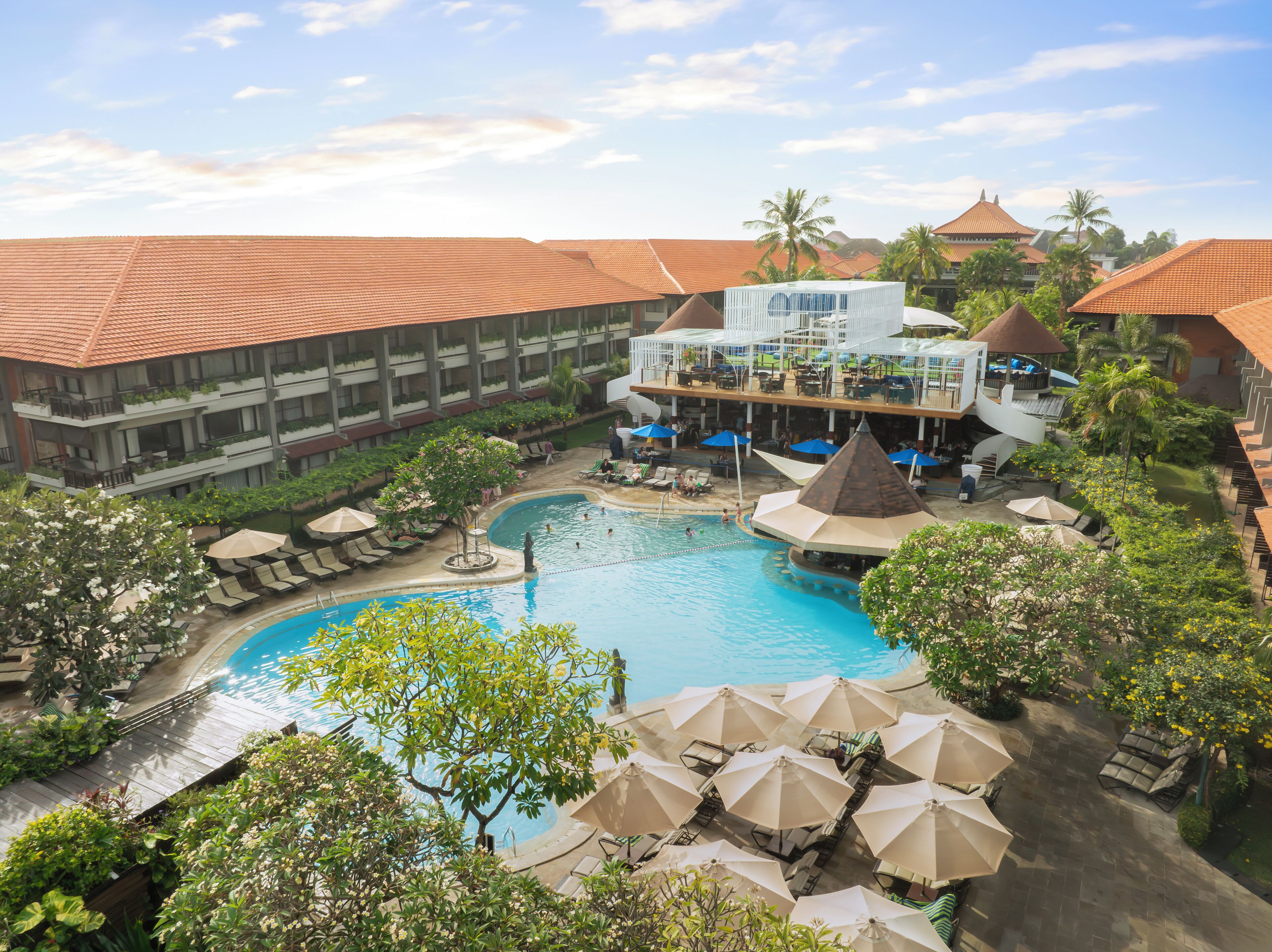 A resort with a swimming pool, lounge chairs, umbrellas, and a multi-story building with balconies.