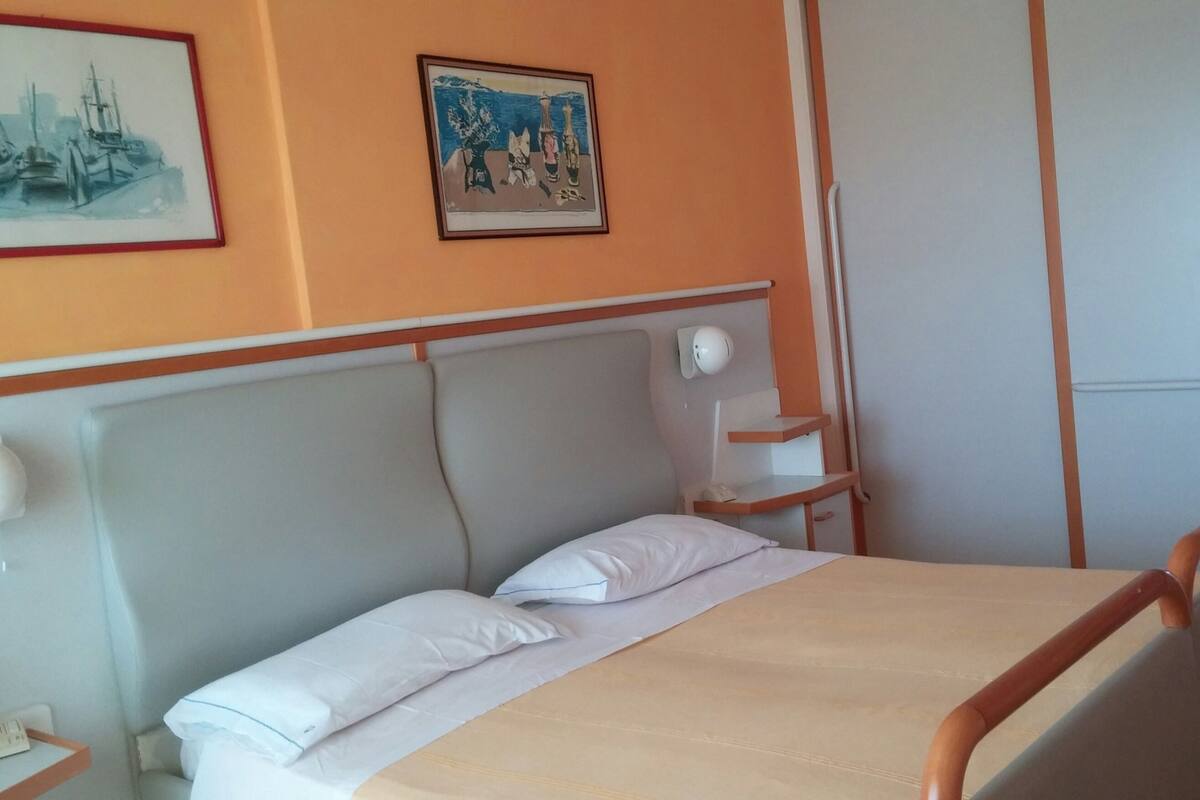 Ein ordentlich bezogenes Bett mit Holz Kopfteil, ein Nachttisch mit Lampe und ein gerahmtes Bild an der Wand.