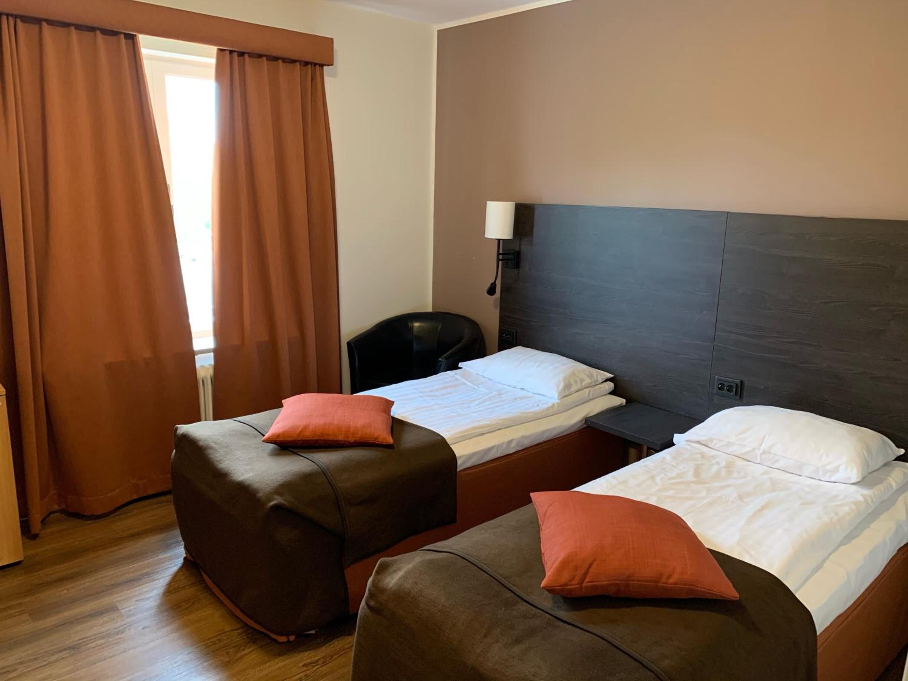Quarto de hotel com duas camas, cada uma com travesseiro e cabeceira. Há uma cadeira e um abajur na parede.