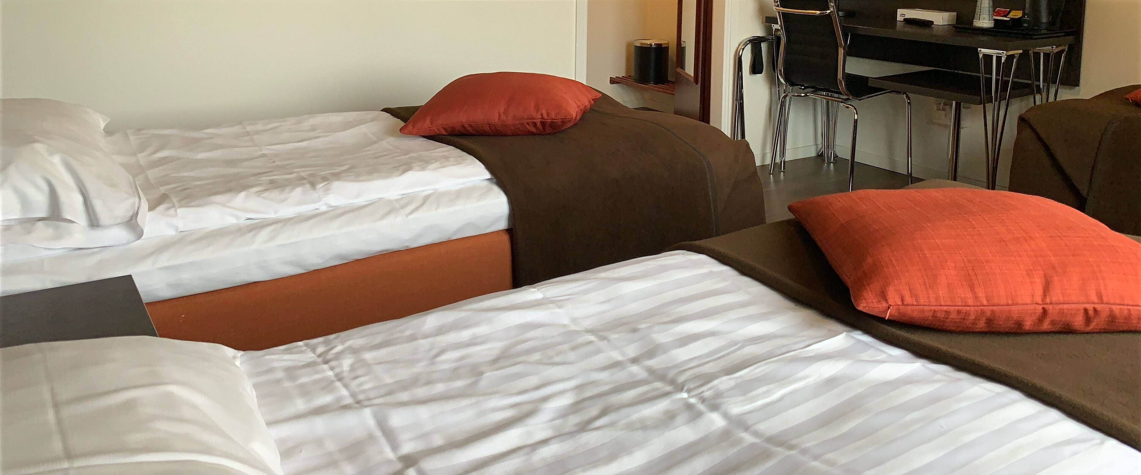 A hotel room with two beds, each with white linens and red pillows. There is a small desk with a chair and a trash can.