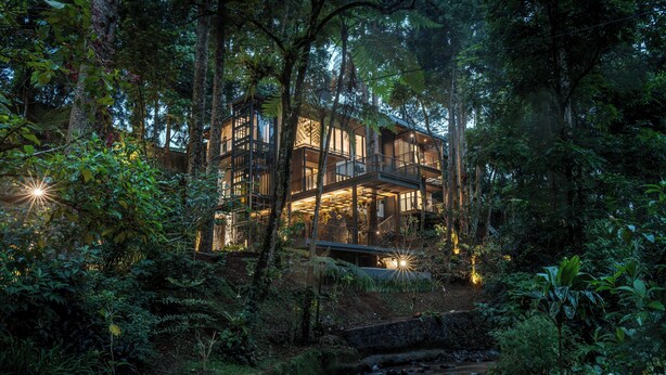 A modern building with a glass facade, surrounded by dense forest and a stream. - Plataran Puncak Residences (Cisarua)