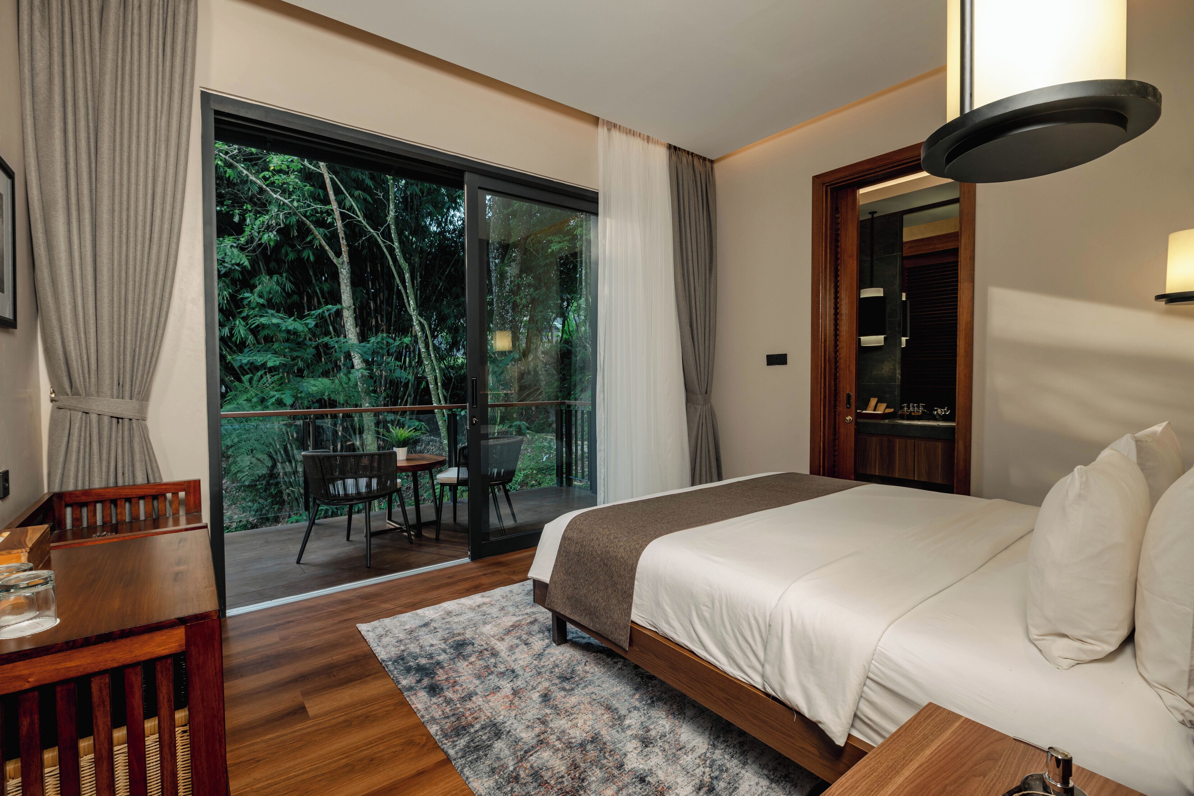 A bedroom with a large bed, a wooden desk, a chair, and a view of a balcony with a table and chairs.