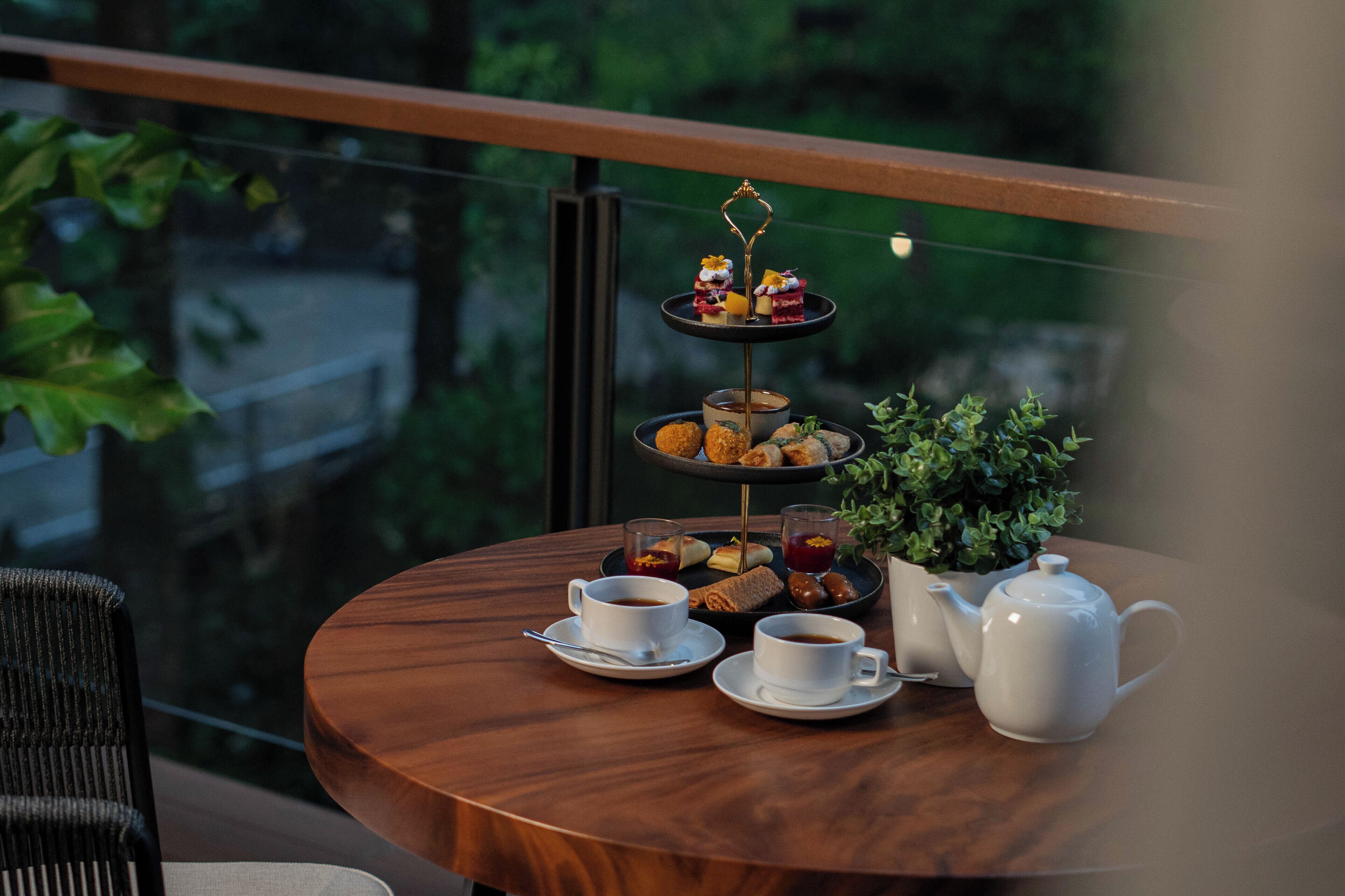 A round wooden table with a tiered dessert stand, two cups of tea, a teapot, and a small potted plant.