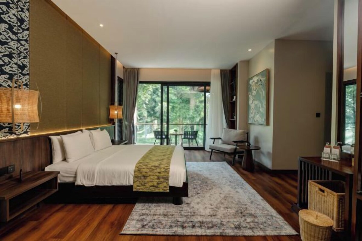 A modern hotel room with a large bed, a sitting area with a table and chairs, and a balcony with a view of the outdoors.