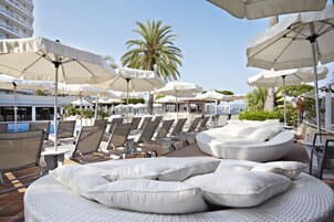 Un área de descanso en la azotea con sillones blancos, sombrillas, una piscina y palmeras.