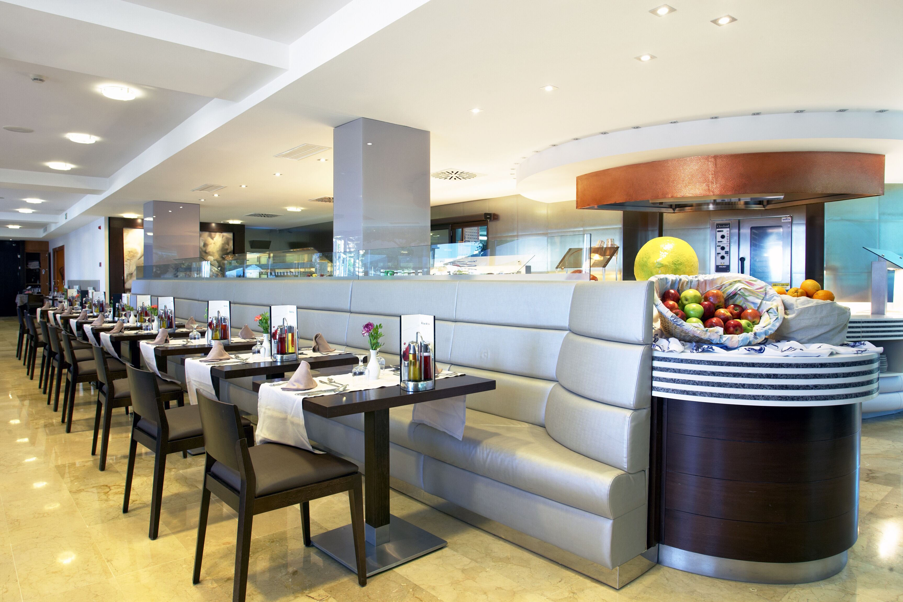 Un comedor moderno con varias mesas y sillas, un área de estar central y una exhibición de frutas.