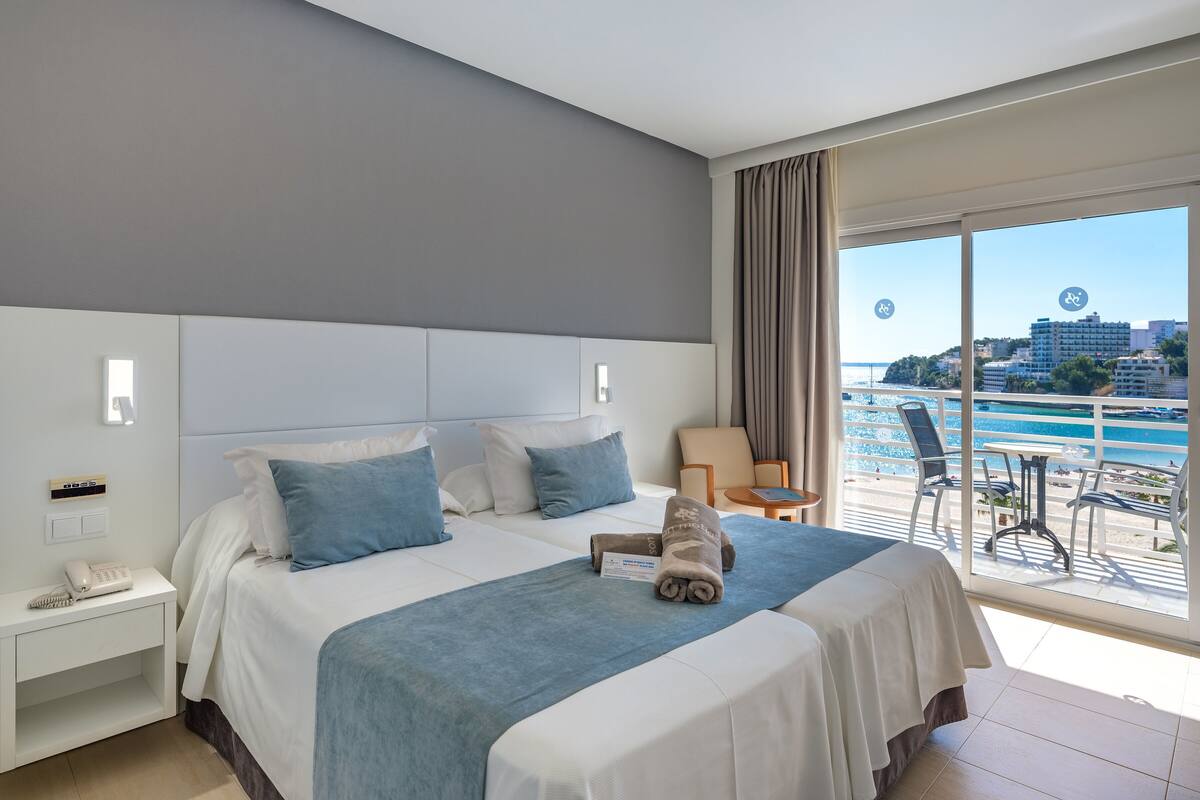 Ein Hotelzimmer mit einem großen Bett, einem Nachttisch mit Telefon, einem kleinen Tisch mit Handtuch und einem Balkon mit Blick auf das Meer und Gebäude.