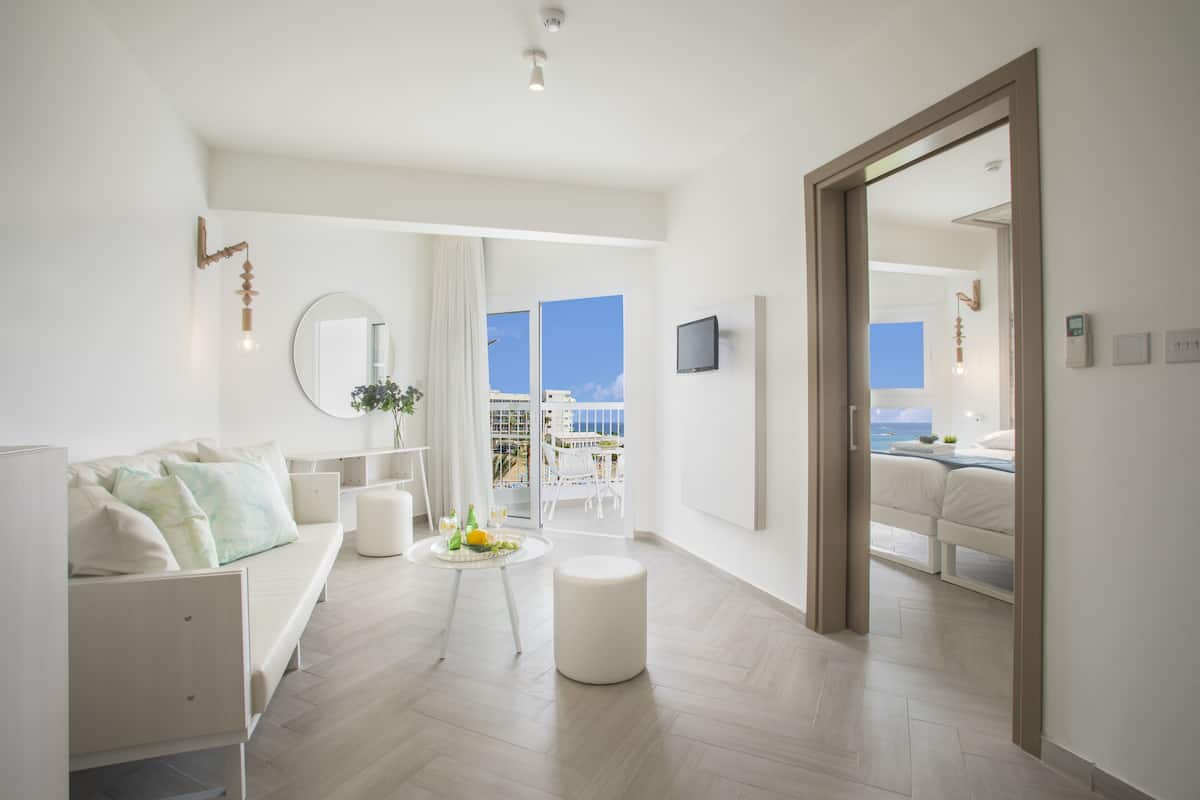 A modern, well-lit living room with a sofa, a round mirror, a small table with a plant, and a view of a balcony with outdoor furniture.