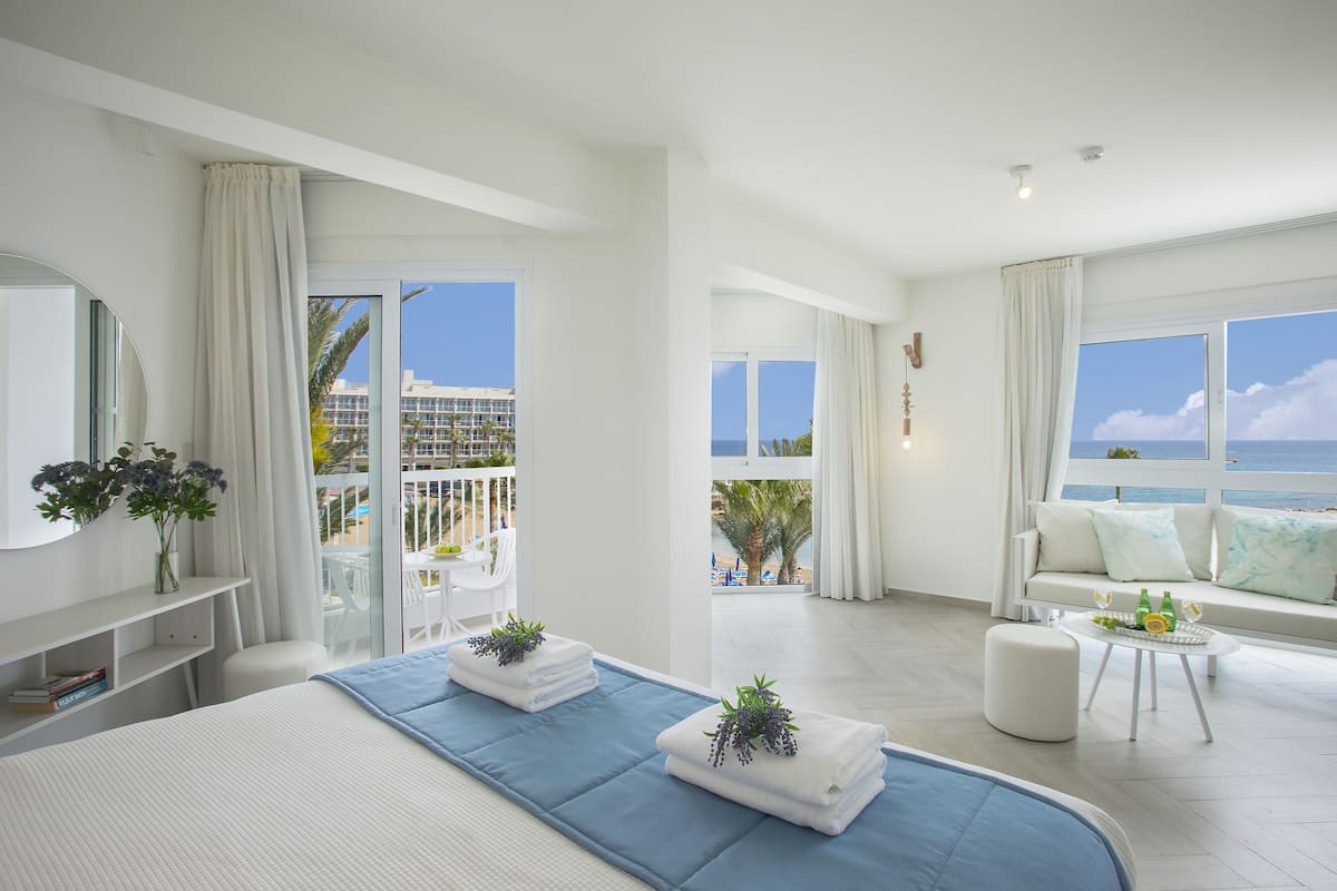 A modern hotel room with a large bed, a balcony with a view of the sea, a sofa, and a small table with drinks.
