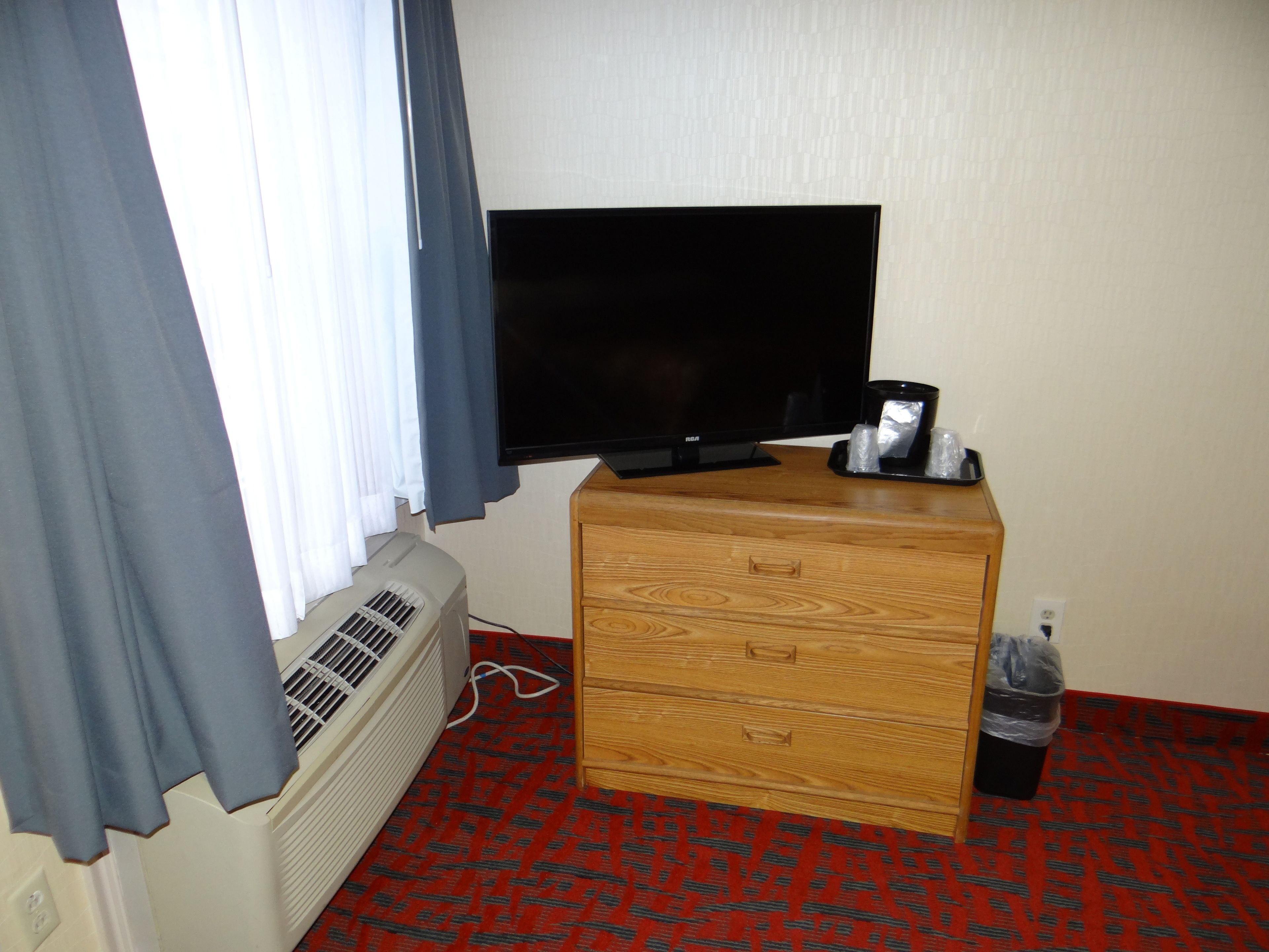 A hotel room with a flat-screen TV on a wooden dresser, a window with curtains, a wall-mounted heater, and a trash bin.