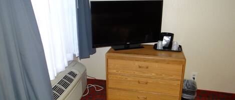 A hotel room with a flat-screen TV on a wooden dresser, a window with curtains, a wall-mounted heater, and a trash bin.