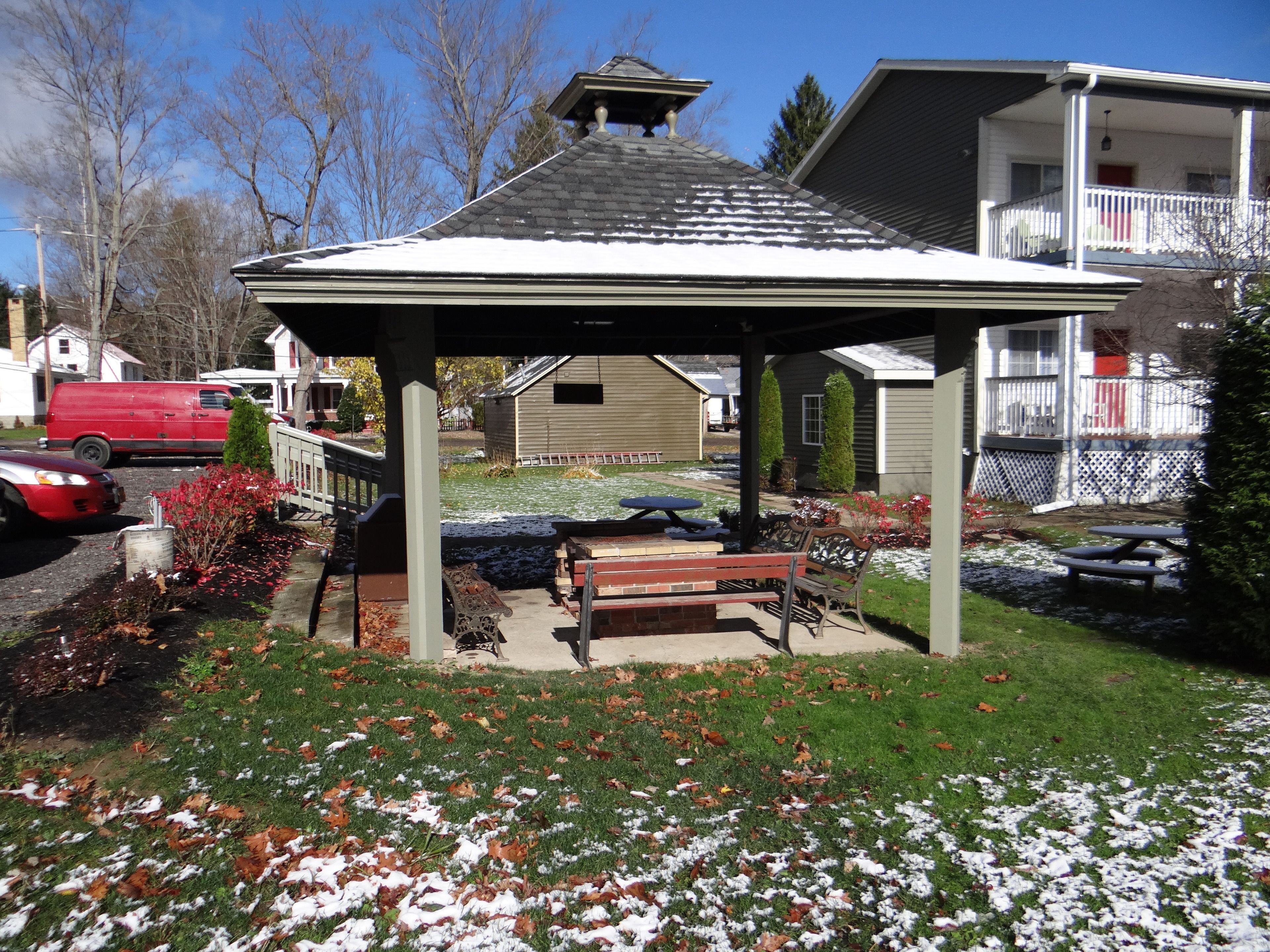 A gazebo with a bench, surrounded by a garden with snow patches and fallen leaves.