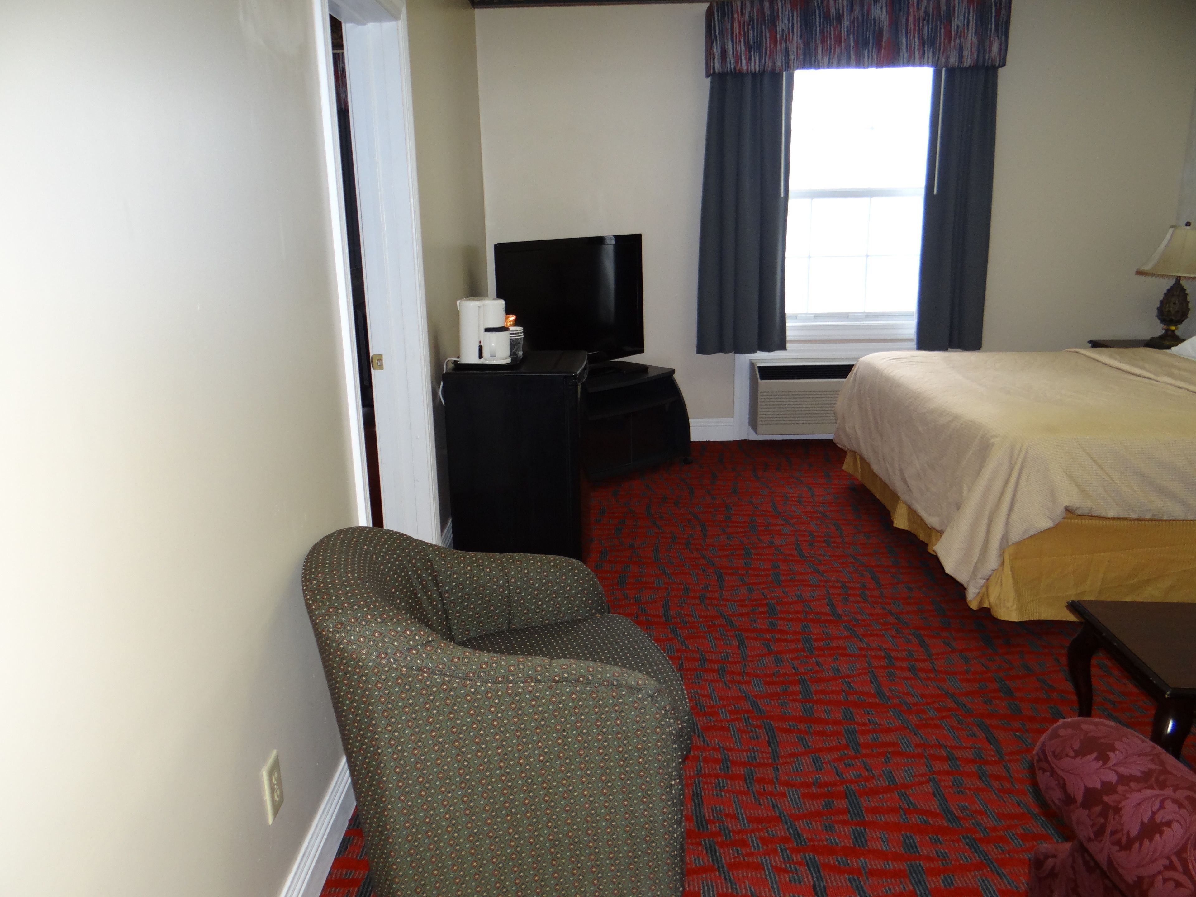 A hotel room with a bed, a television on a stand, a chair, a small table, and a window with curtains.