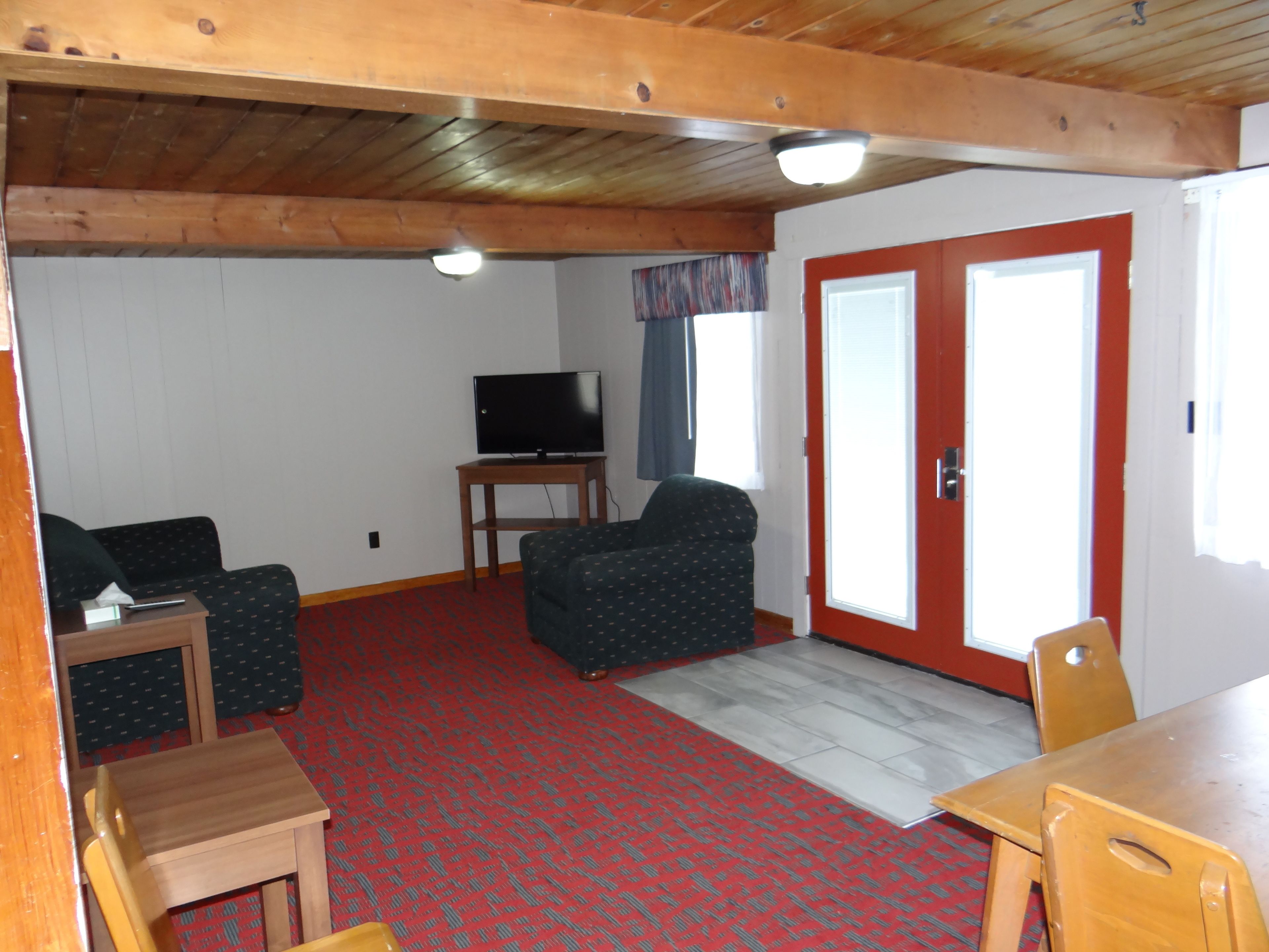 A living room with a television, a sofa, a chair, and a table. There is a red carpet and a wooden ceiling.