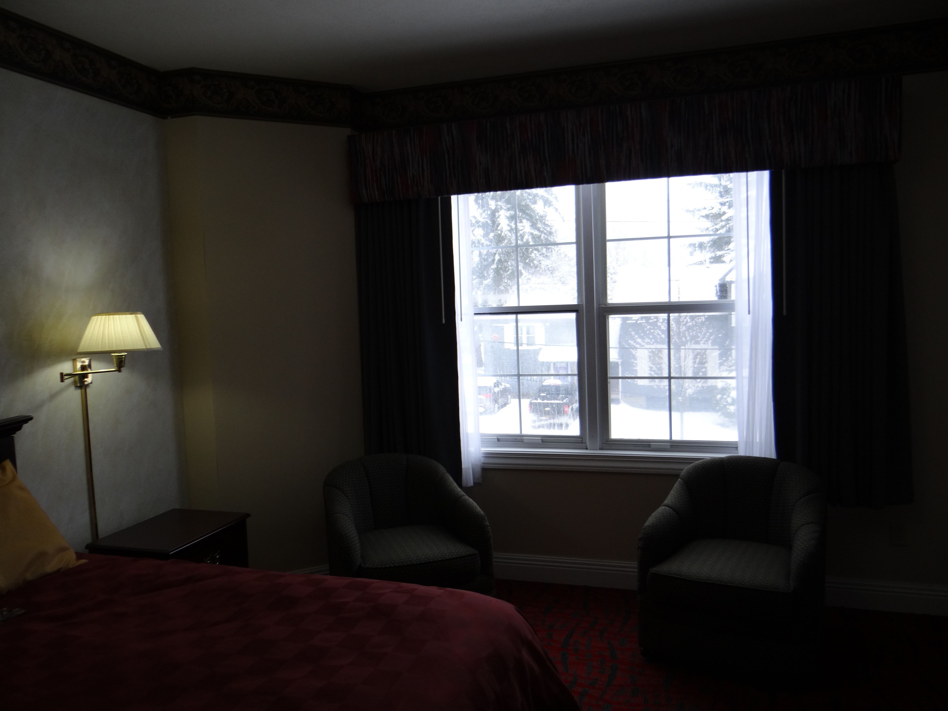 Ein Hotelzimmer mit großem Fenster, zwei Sesseln, einem Bett mit roter Bettdecke, einem Nachttisch und einer Stehlampe.