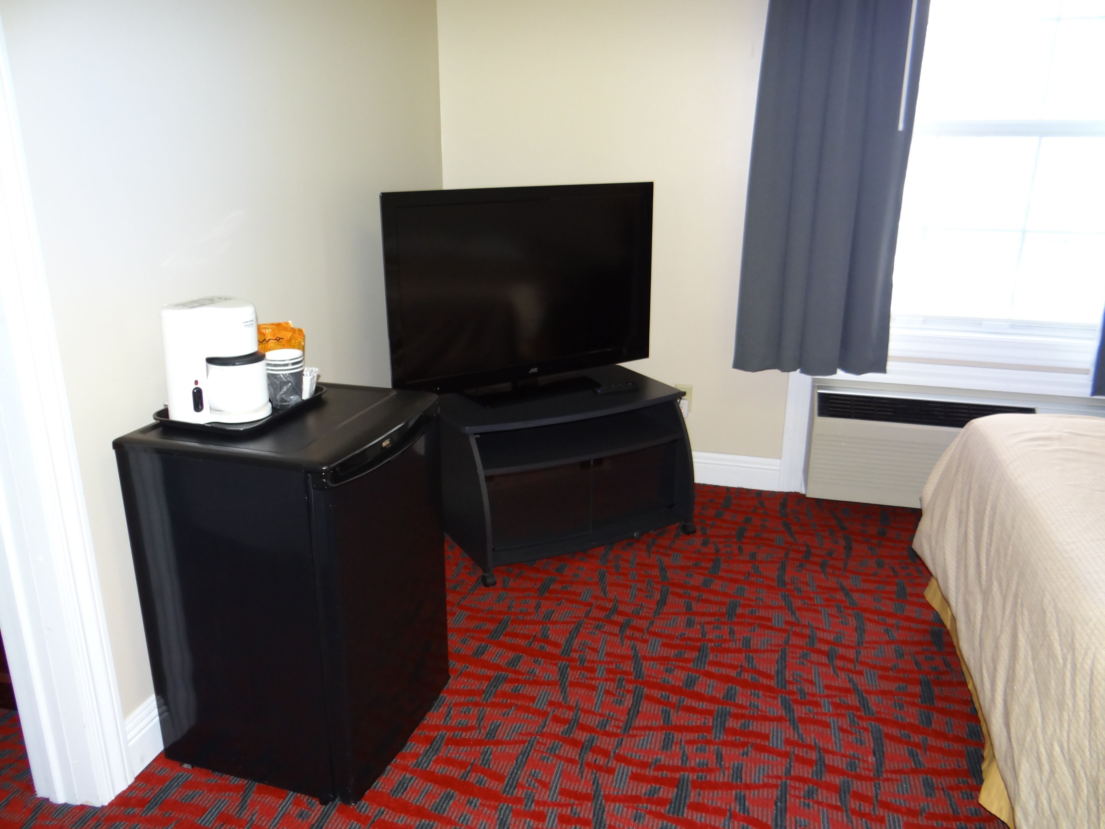 Habitación de hotel con televisor de pantalla plana sobre un soporte negro, cafetera y cama con alfombra roja estampada.