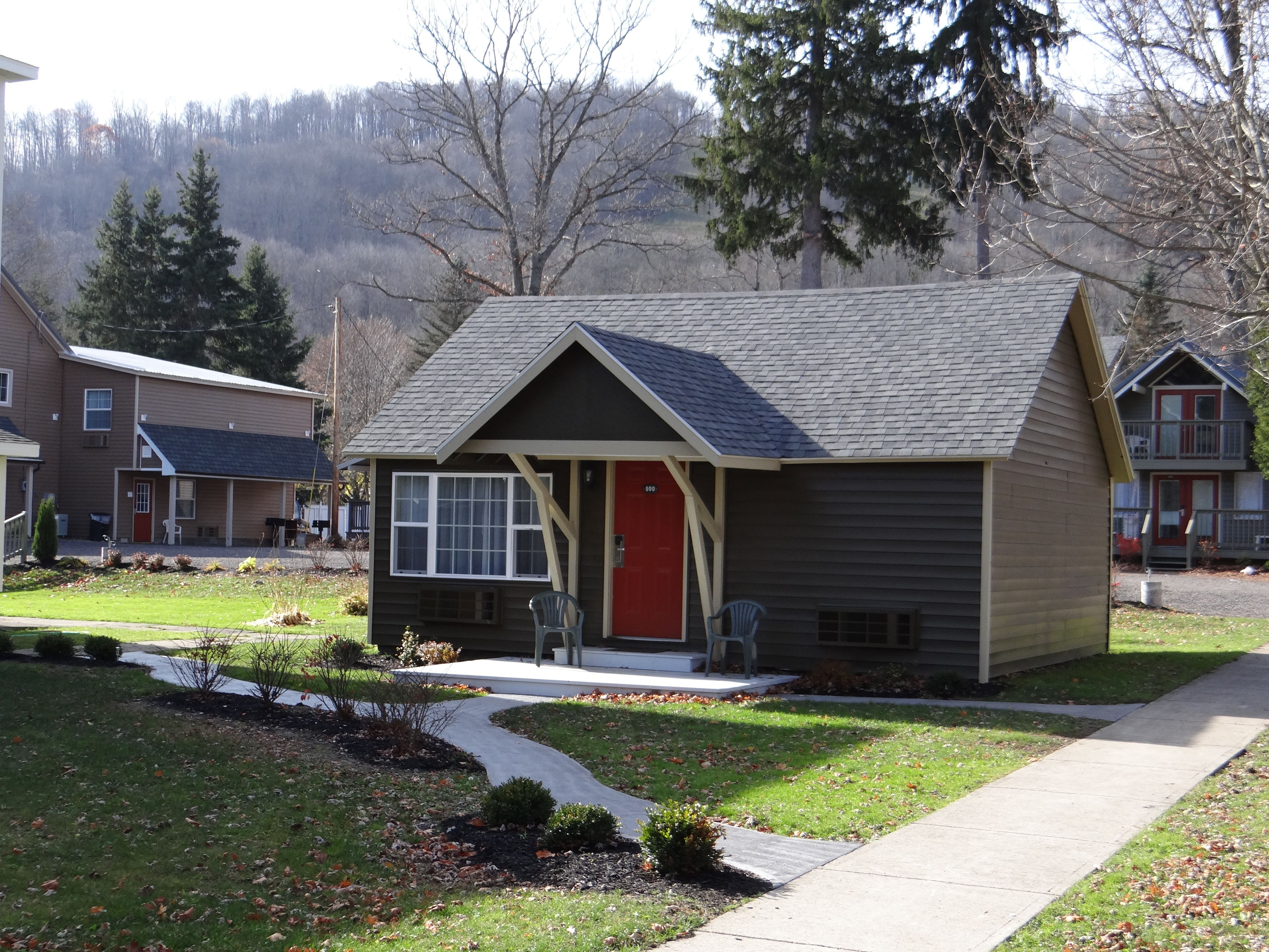 A small, single-story house with a red front door, a covered porch, and a small front yard with a pathway leading to the entrance.