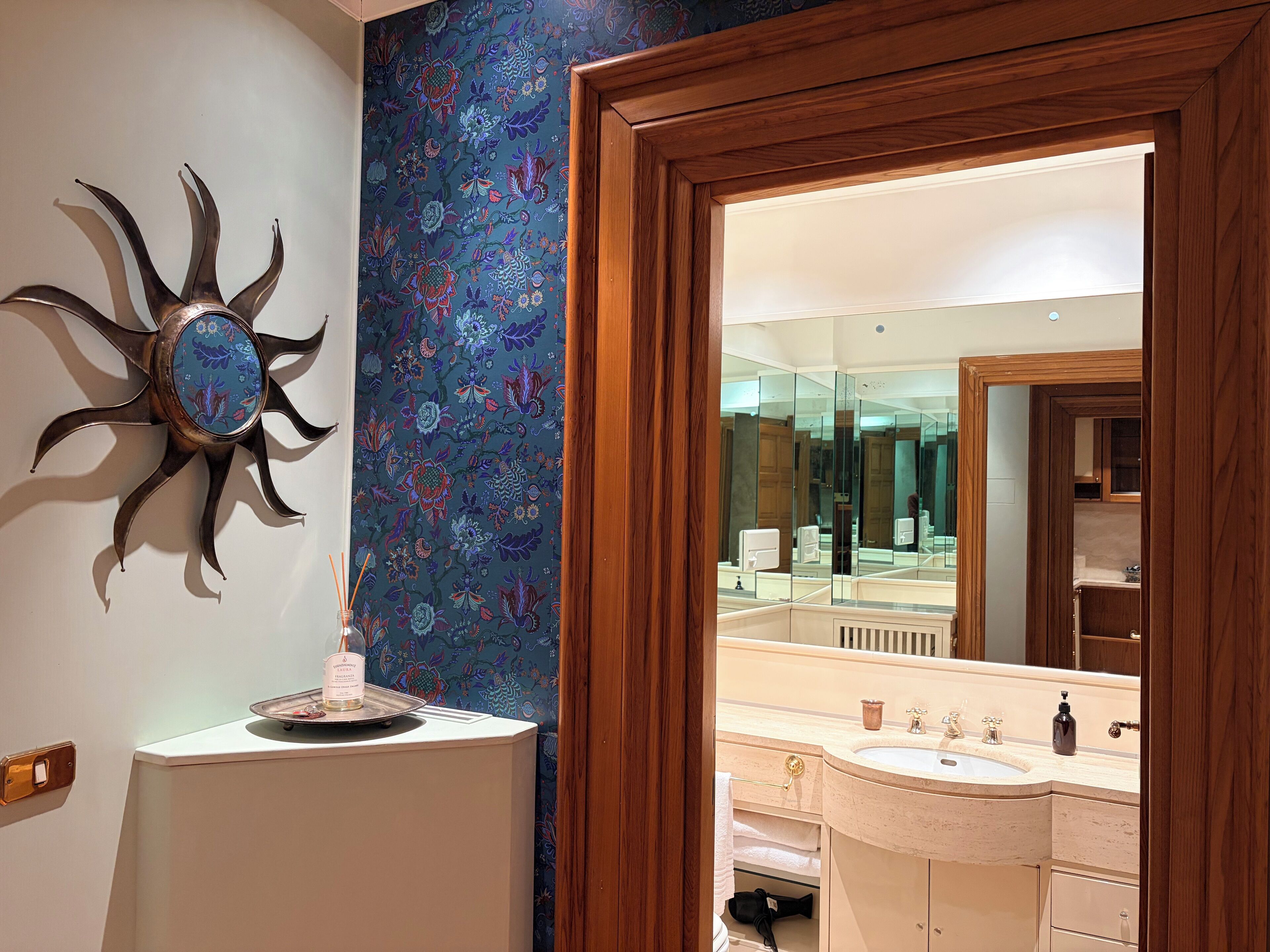 Een badkamer met een grote spiegel, een decoratief zonvormig object aan de muur, en een bloemenpatroon op de muur.