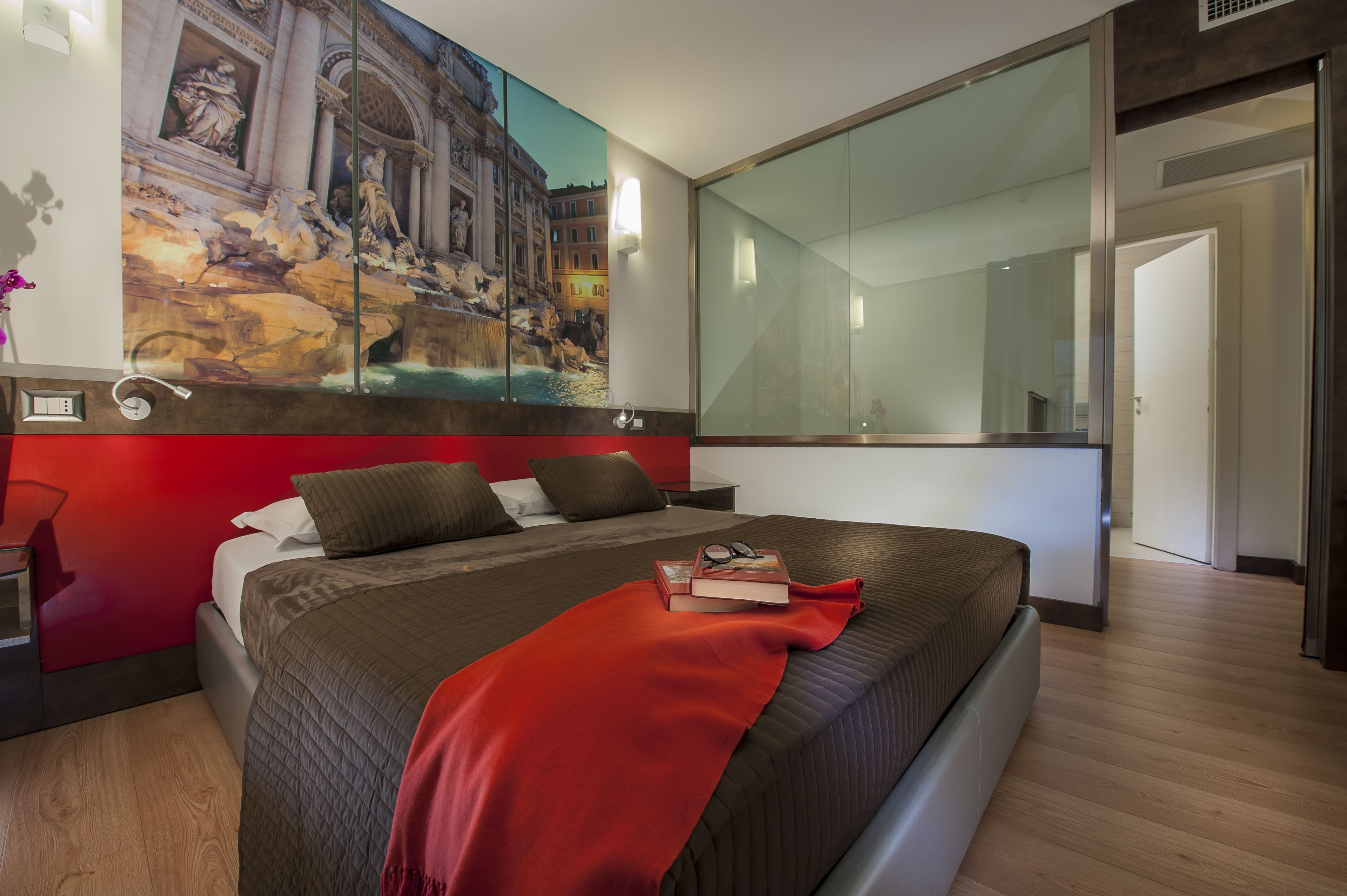 Ein modernes Schlafzimmer mit einem großen Bett, einer roten Tagesdecke, einem Nachttisch, einem Spiegel und einem Gemälde eines historischen Gebäudes.