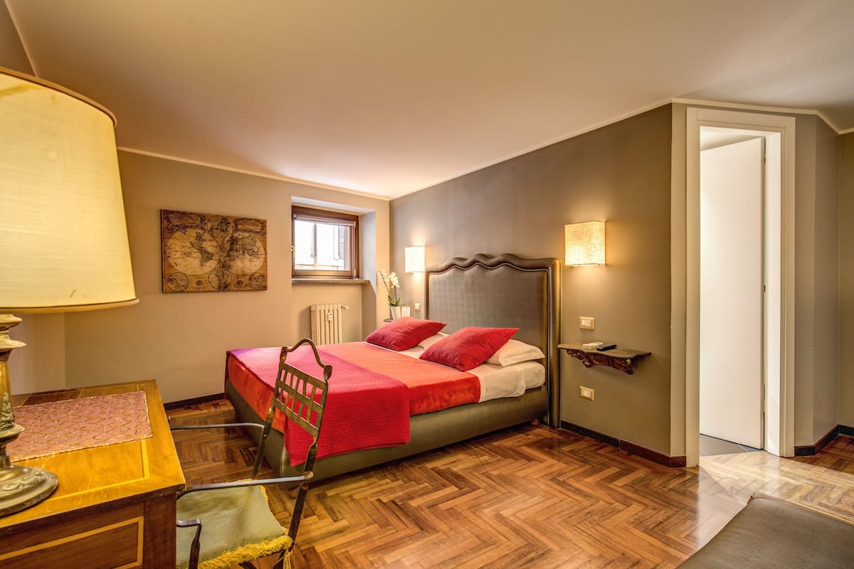 Un dormitorio con una cama, una silla, una mesa, una lámpara y una ventana.