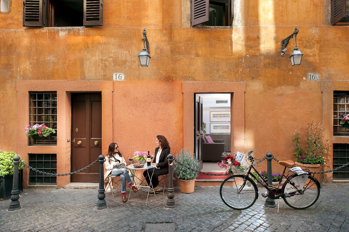 A building with a rustic facade, numbered 16A, featuring a door and windows with shutters, a small outdoor seating area with a table and chairs, and a bicycle parked outside.
