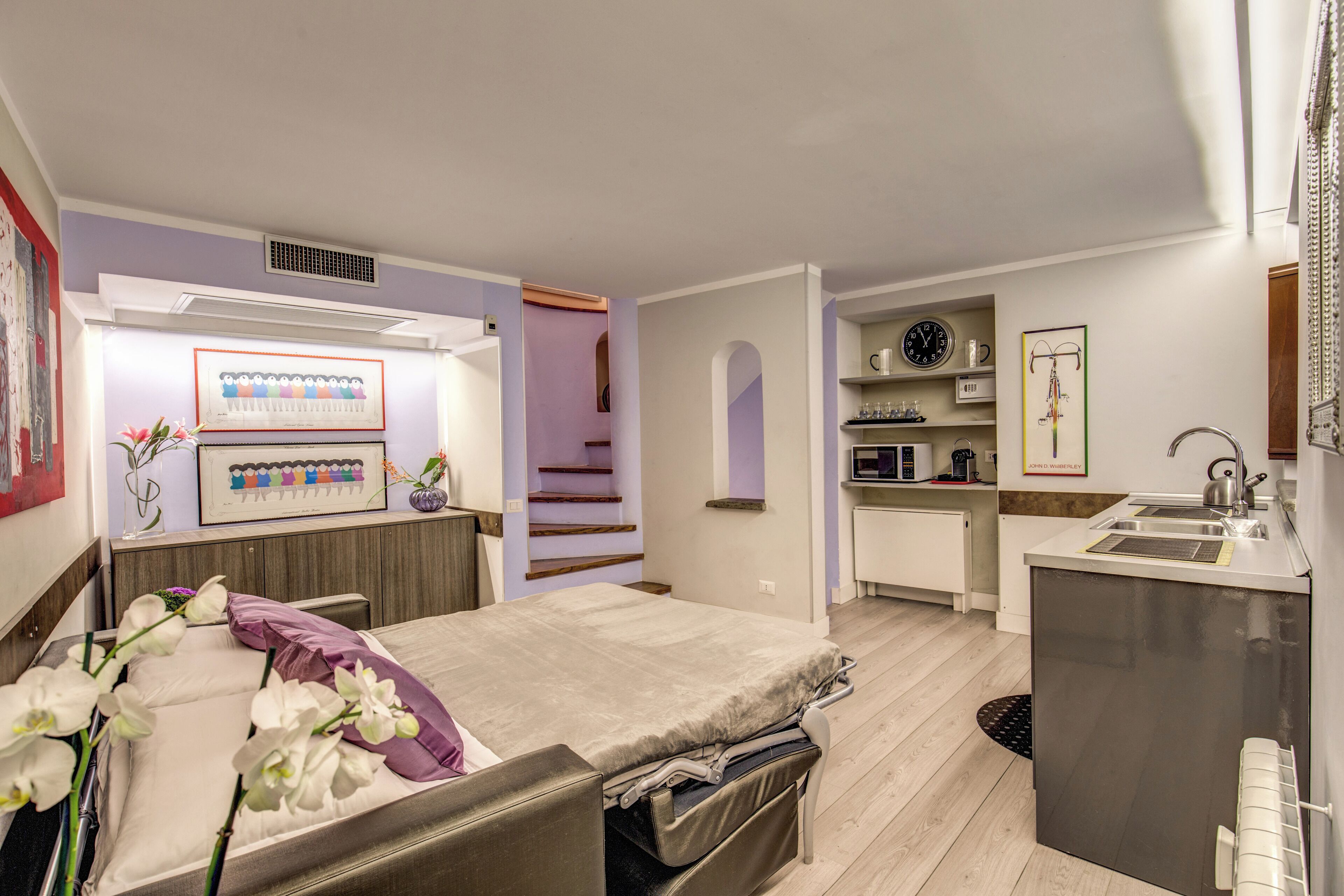 A compact hotel room with a bed, a small kitchenette, a bathroom, and a view of the hallway.