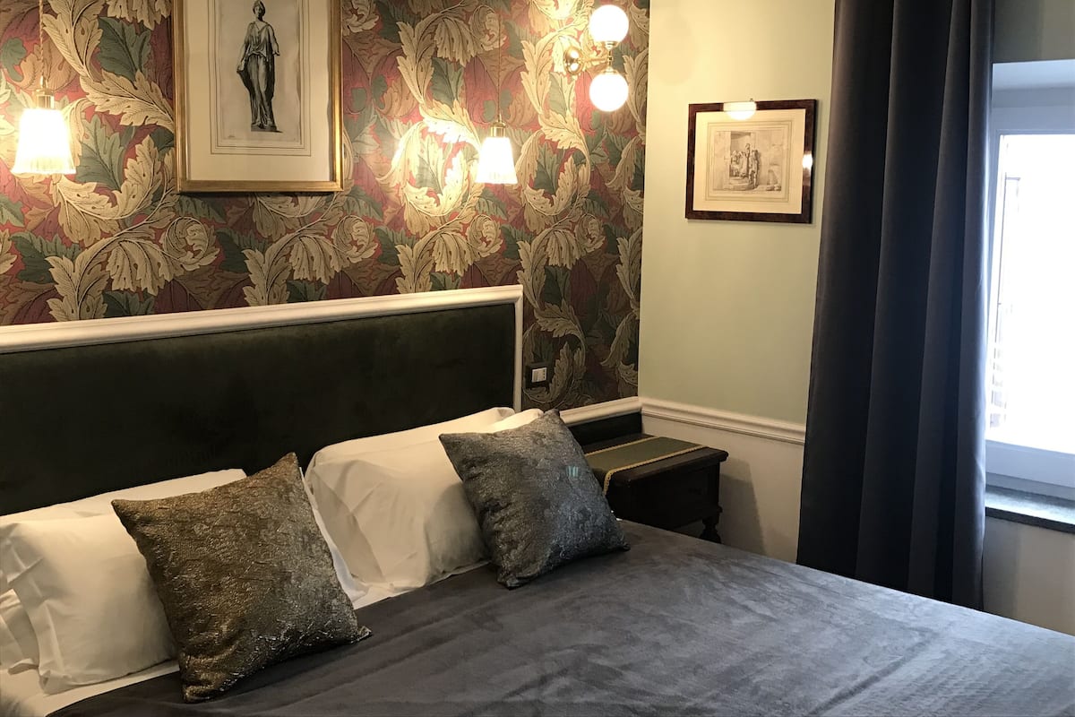 Una cama bien tendida con un cabecero oscuro, dos almohadas y una colcha de color claro. Se ven dos lámparas fijadas a la pared y una obra de arte enmarcada sobre la cama. A la derecha, hay una ventana con cortinas oscuras.