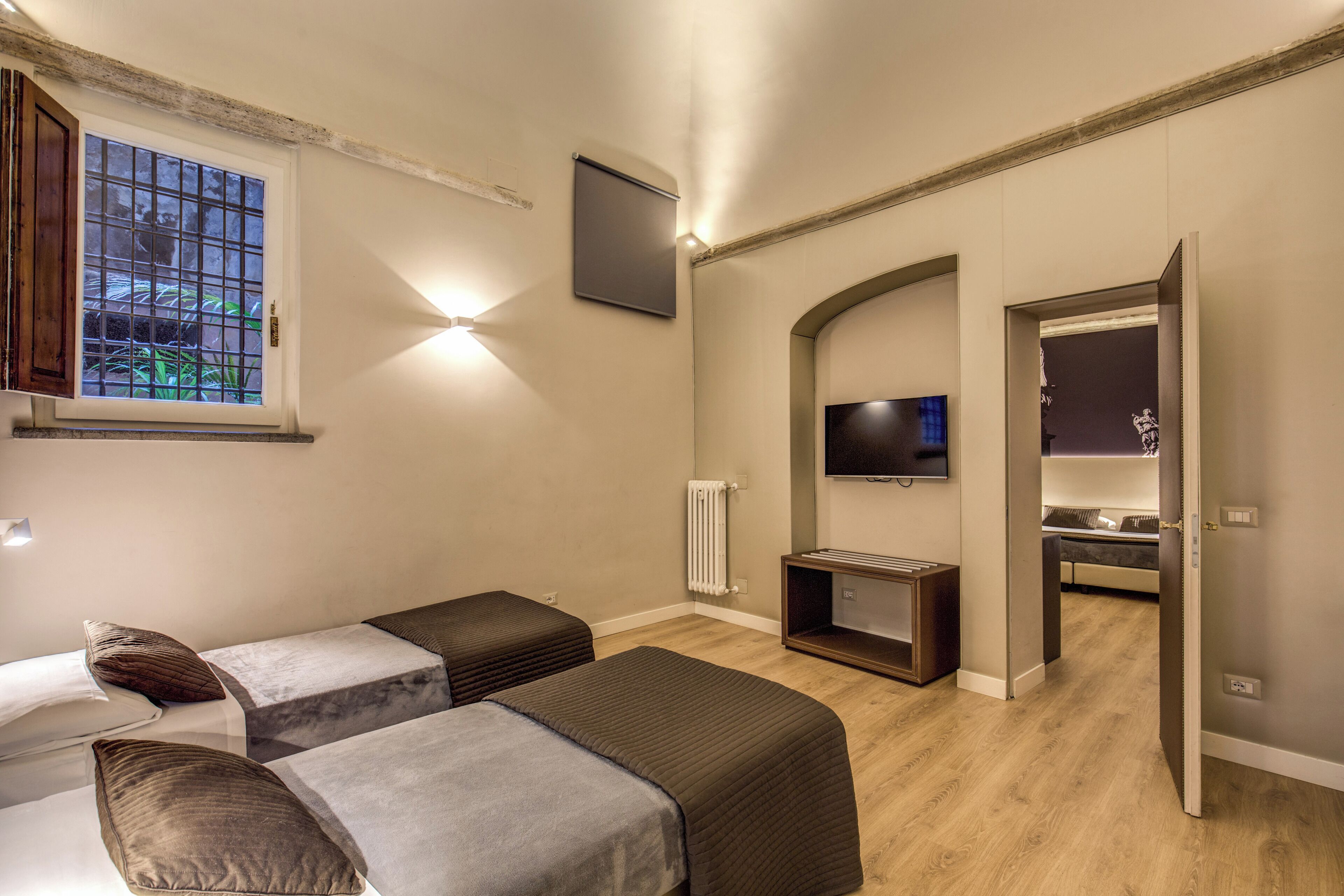 A hotel room with two single beds, a TV, a radiator, and a view of the outdoors through a window.