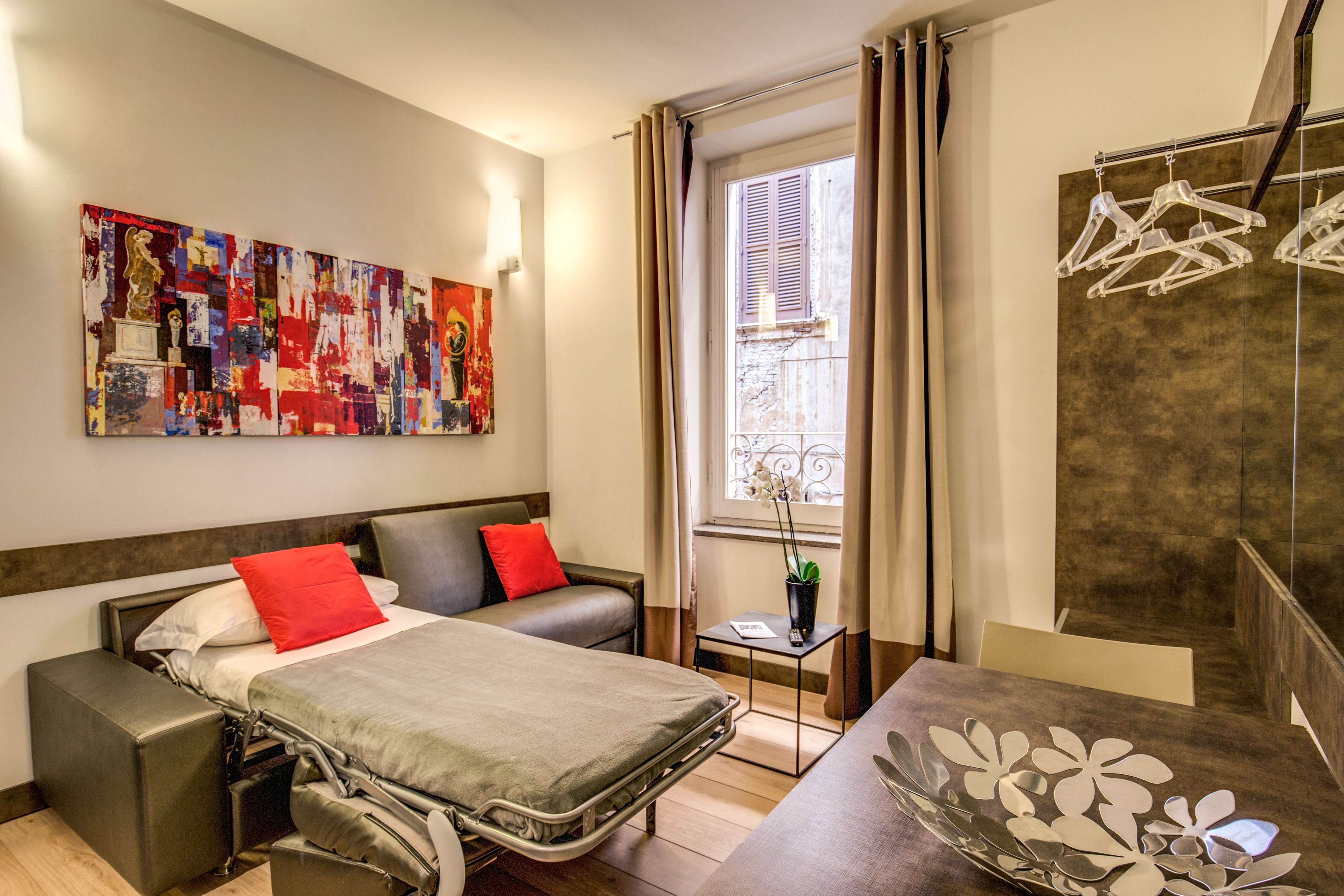 A modern hotel room with a bed, a small table, a chair, a window with curtains, and a wall with artwork.