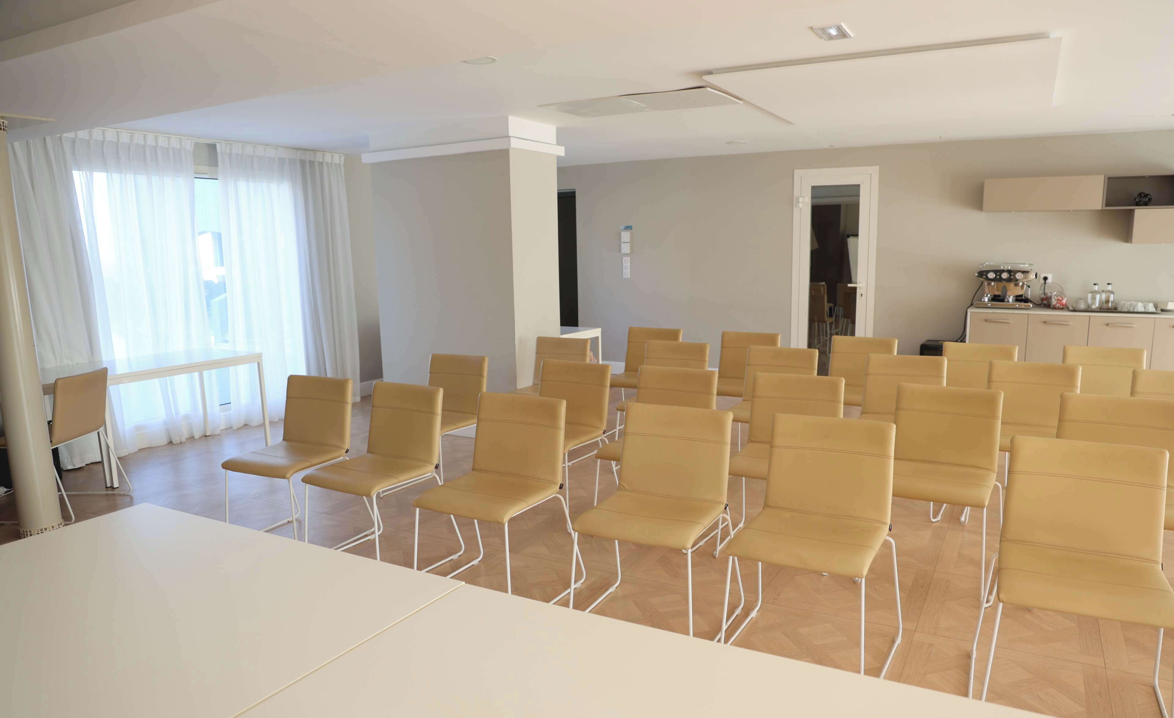 Une salle de conférence avec une longue table et de nombreuses chaises, un coin cuisine et une fenêtre avec des rideaux.