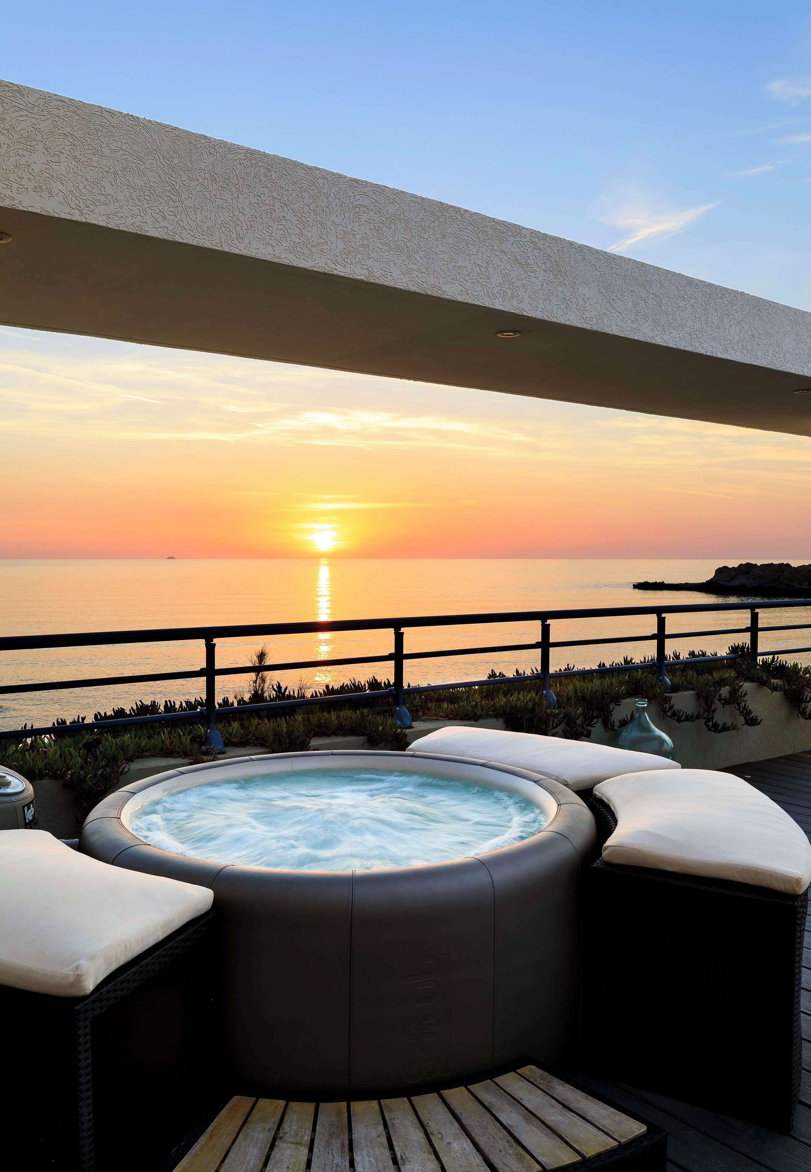 A hot tub with cushions, a wooden table, and a view of the sunset over the ocean.
