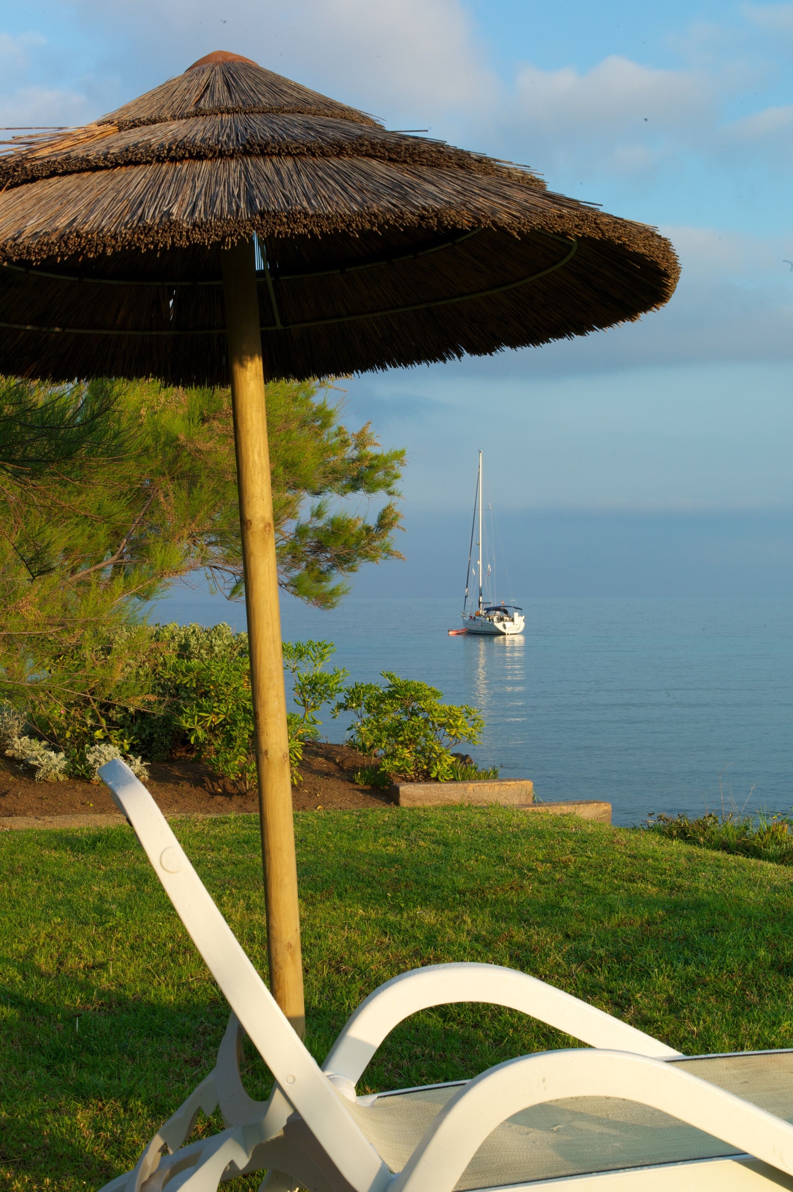 A beachside view with a thatched umbrella, a sailboat, and a white lounge chair.