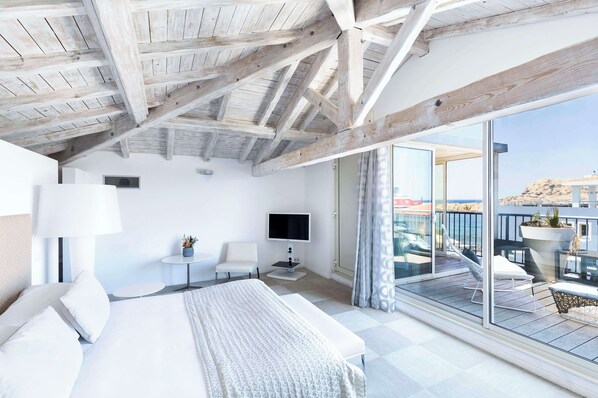 A modern hotel room with a large bed, a TV, a small table with a lamp, and a balcony with a view of the city.