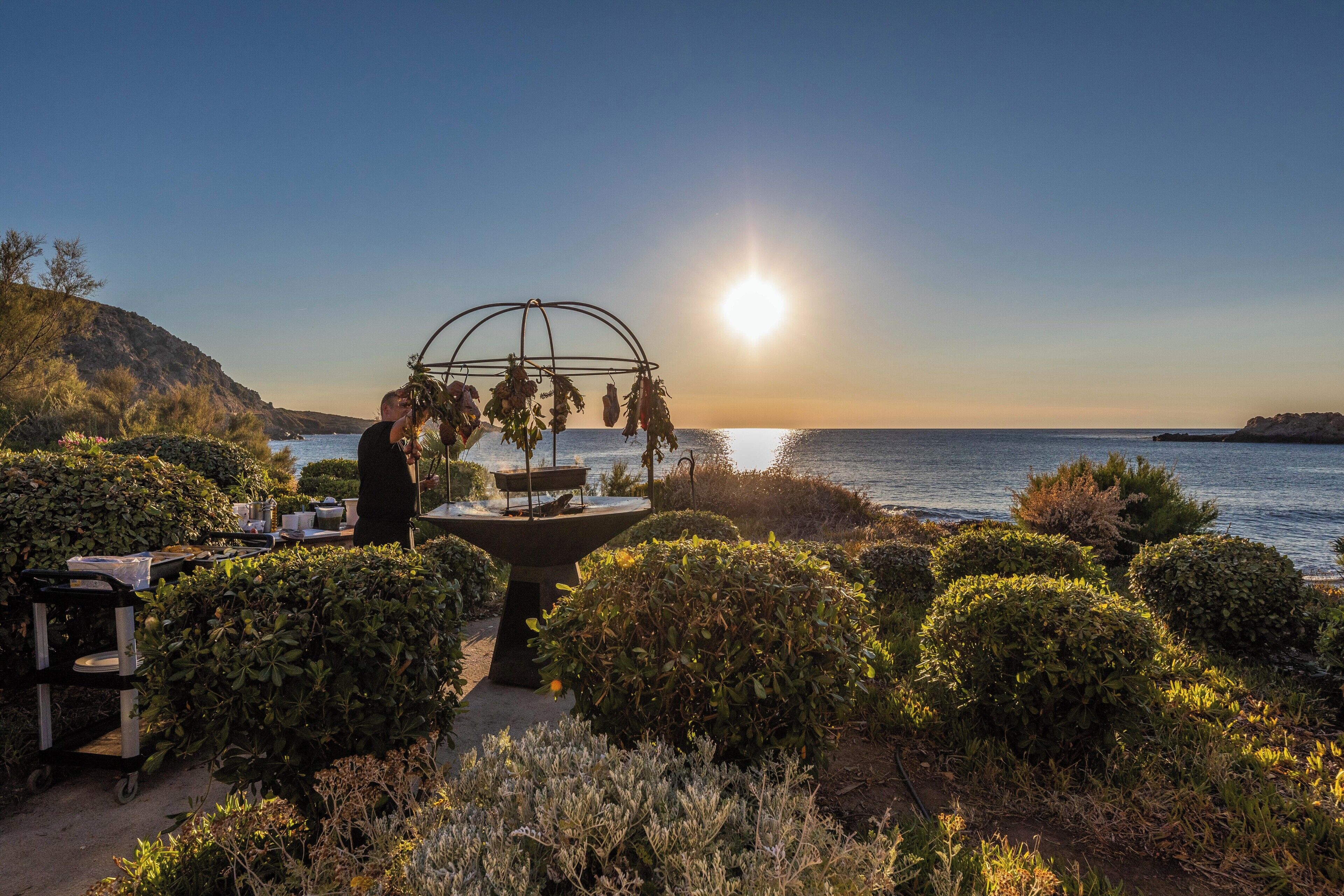 Un jardín con una fuente, una mesa con objetos y vista al mar al atardecer.