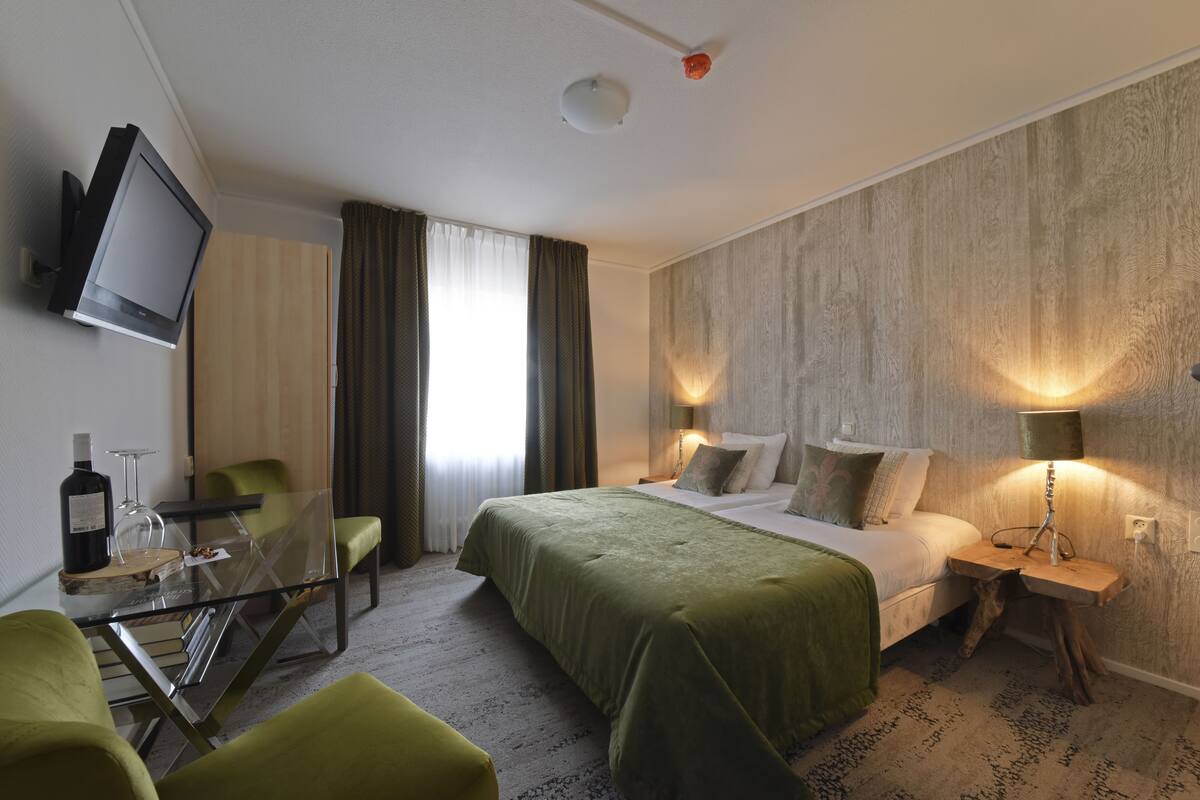 Een hotelkamer met een bed, twee nachtkastjes met lampen, een aan de muur bevestigde televisie, een glazen tafel met een fles en een glas, en een groene stoel.