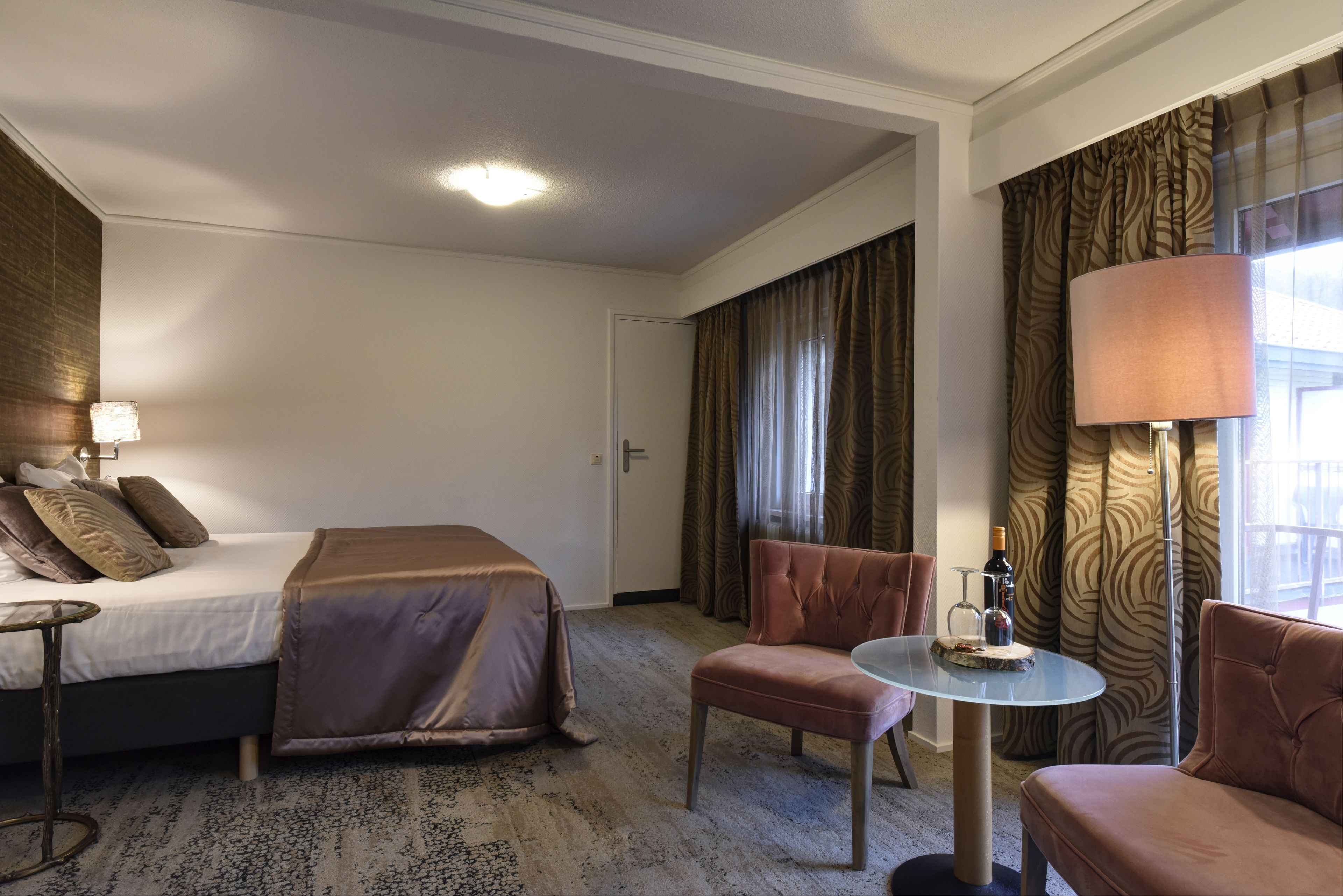 A hotel room with a bed, two armchairs, a small table, a lamp, and a bottle of wine.