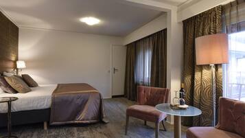 A hotel room with a bed, two armchairs, a small table, a lamp, and a bottle of wine.
