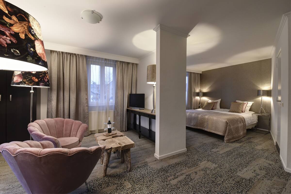 Een hotelkamer met een groot bed, een bureau met televisie, een roze fauteuil en een tafel met een fles.