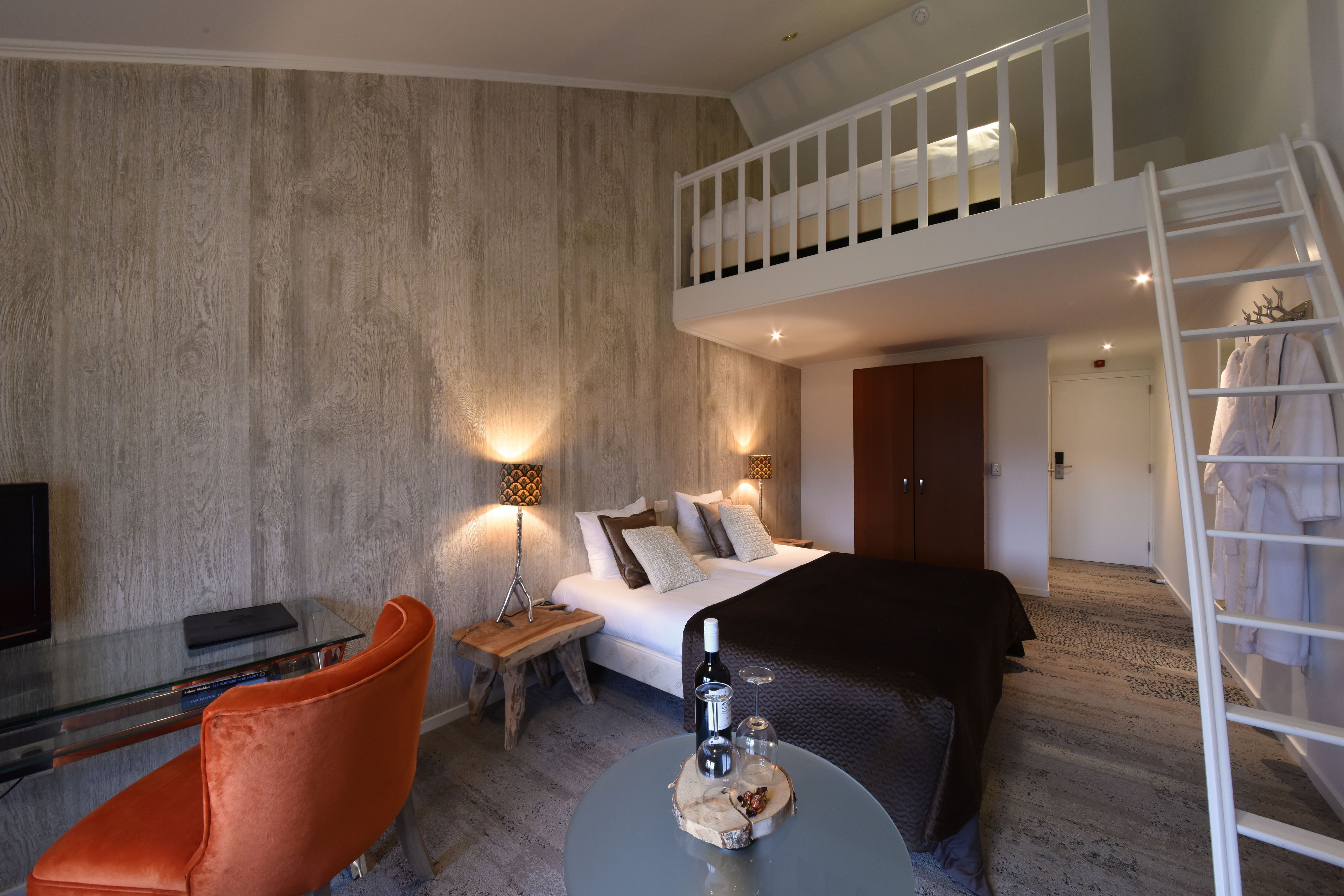Une chambre d’hôtel moderne avec un lit en mezzanine, un bureau en verre, un fauteuil orange confortable et une petite table ronde sur laquelle se trouvent une bouteille et un verre.