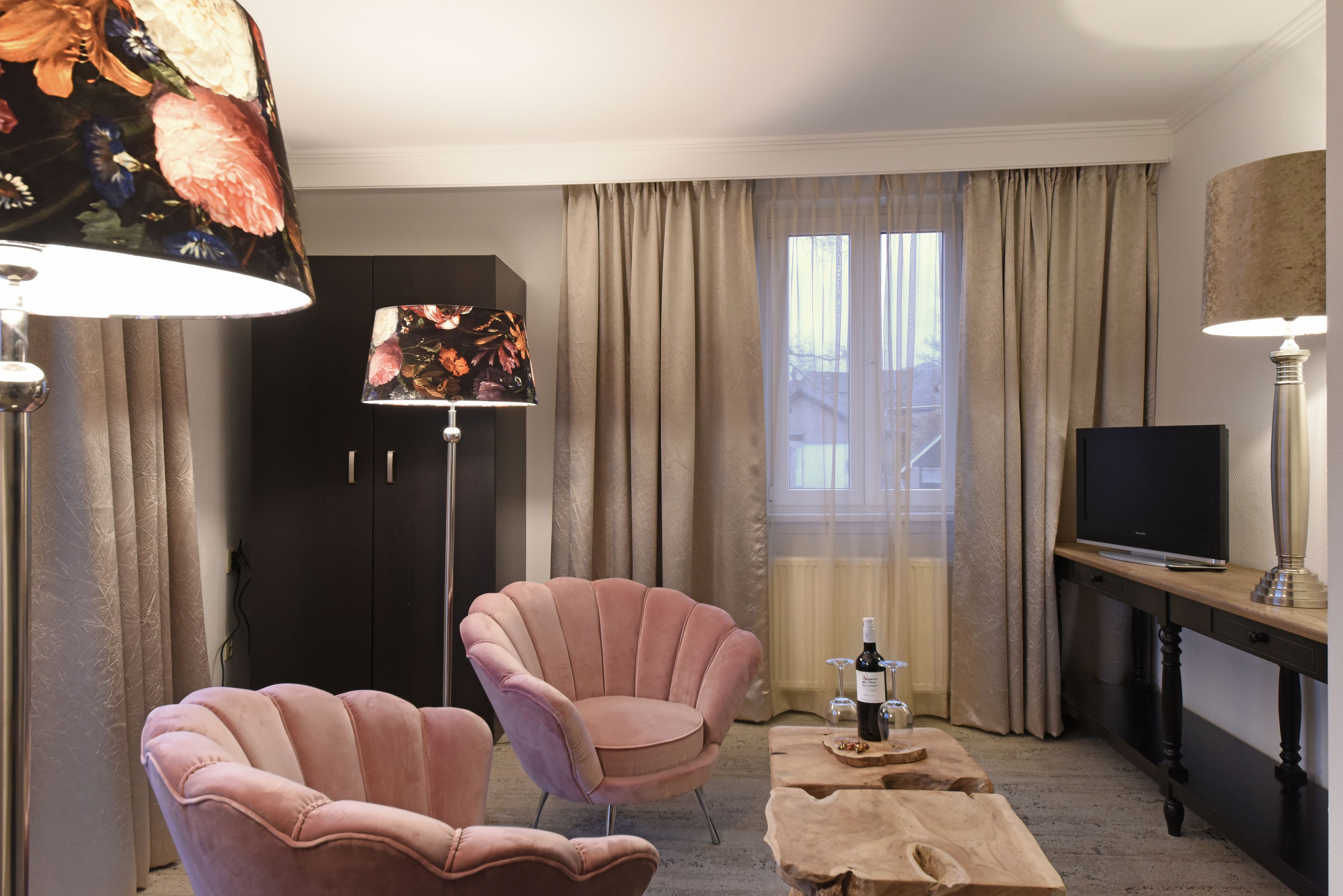 A hotel room with two pink armchairs, a wooden coffee table, a TV, and a lamp with a floral lampshade.