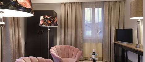 A hotel room with two pink armchairs, a wooden coffee table, a TV, and a lamp with a floral lampshade.