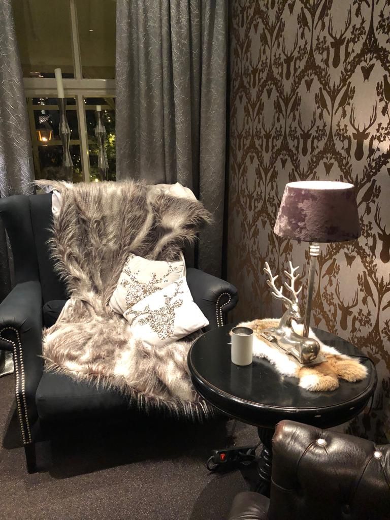 Una habitación con un sillón de cuero negro, un plaid de piel sintética, una almohada blanca, una mesita redonda pequeña con una lámpara y una pared decorativa con un motivo de ciervo.