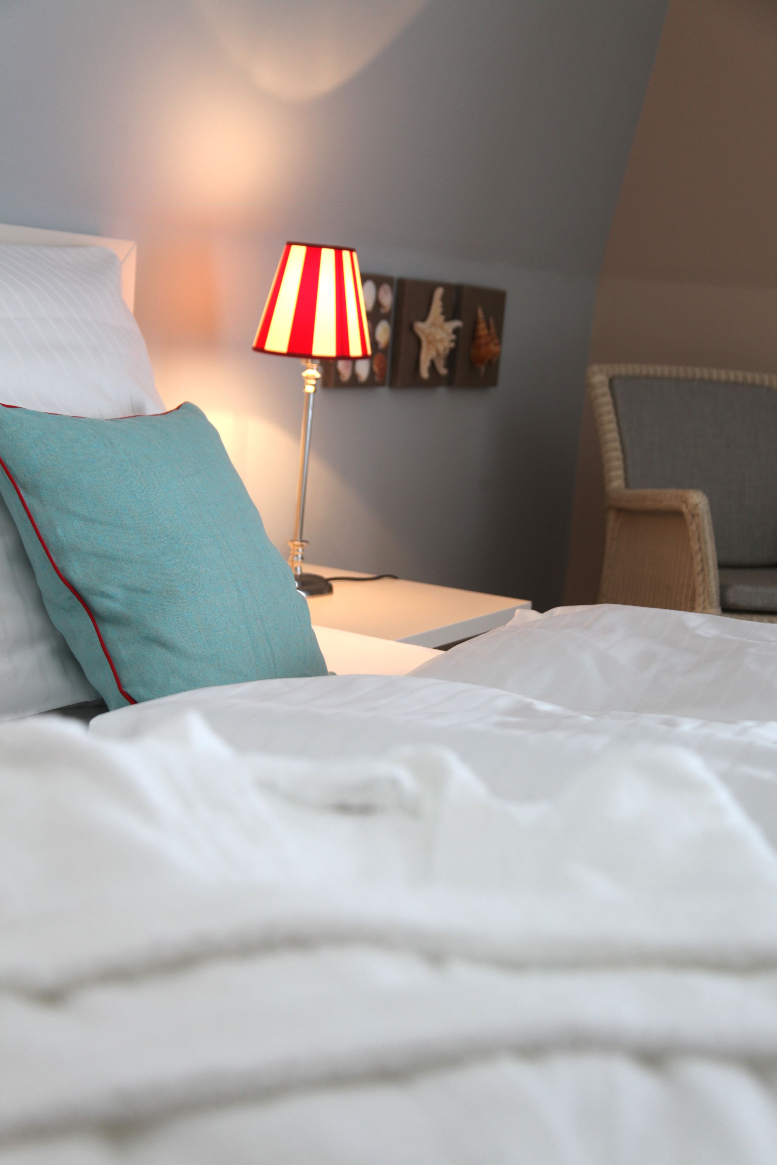 Una cama bien hecha con sábanas blancas y almohadas color turquesa, una lámpara a rayas sobre una mesita de noche y una silla con cojín.