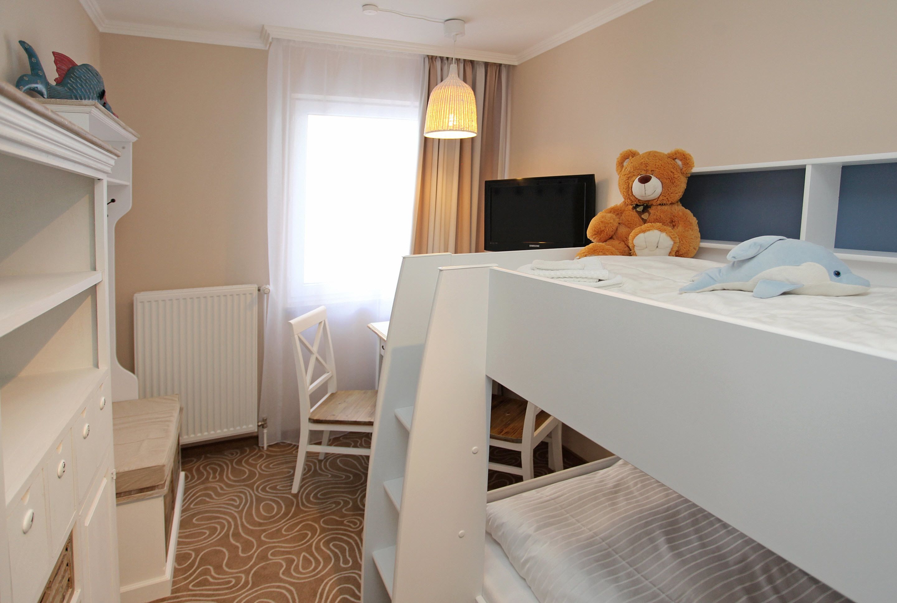 Quarto de criança com beliches, televisão, um urso de pelúcia e uma cadeira.