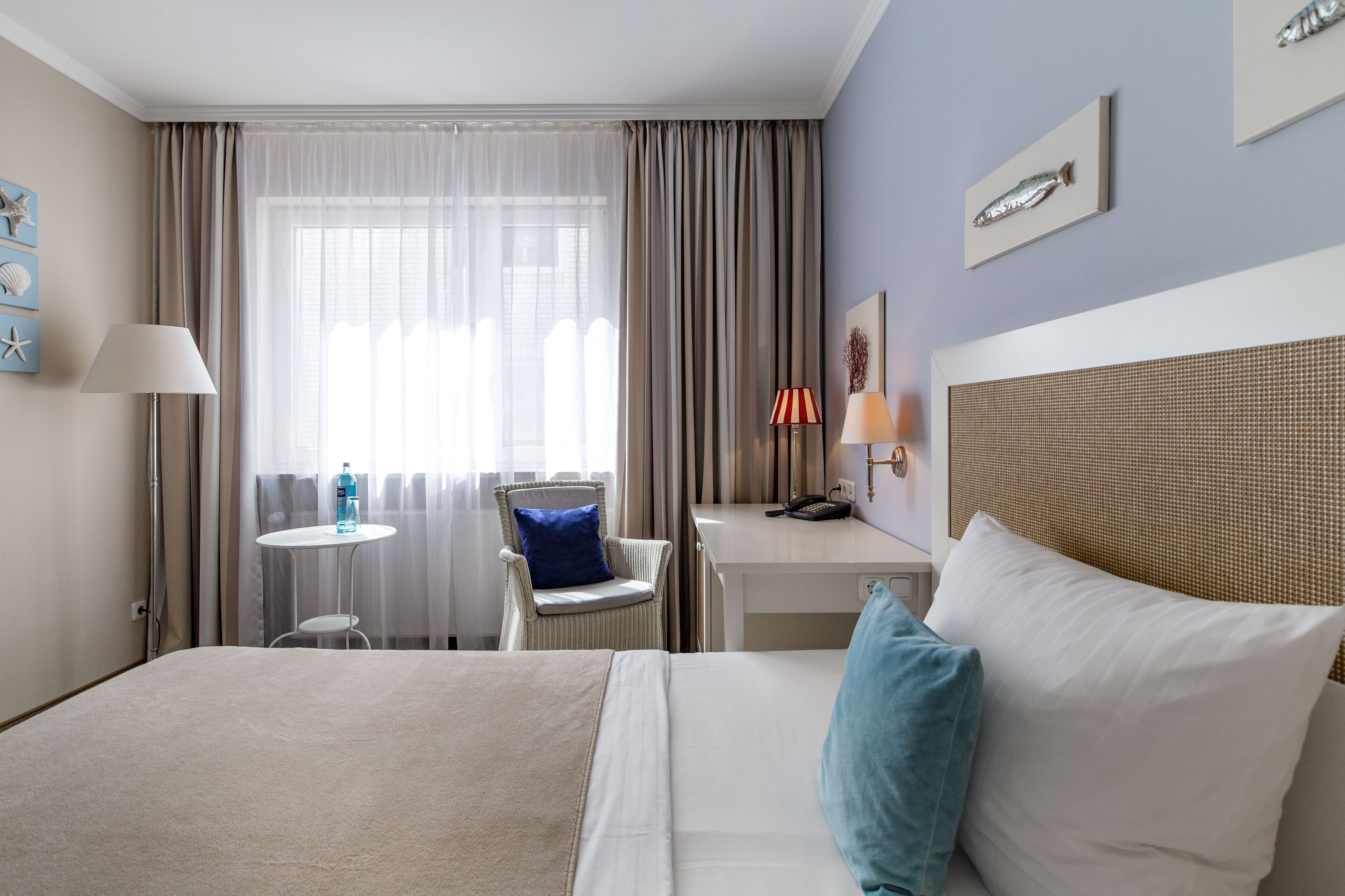 A hotel room with a large bed, a desk, a chair, a small table, and a window with curtains.