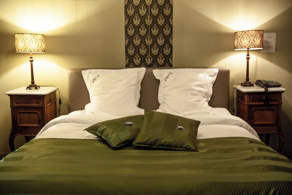 Una cama bien hecha con dos almohadas, un edredón verde y dos mesitas de noche con lámparas.
